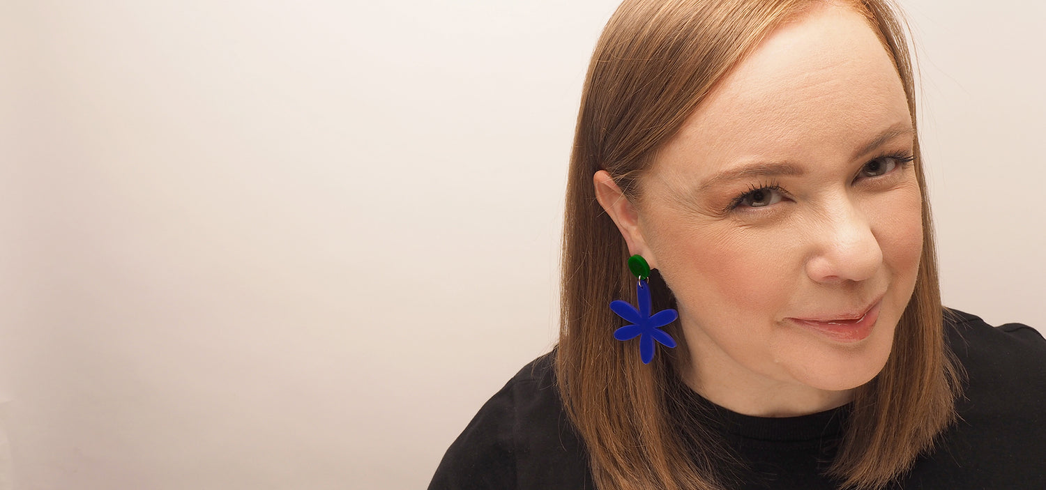 A curvy, red-headed woman wearing the Is Daisy Blue dangle earrings. The earrings are made from a forest green acrylic button and dark blue acrylic daisy dangle. The acrylic pieces are connected with a small, silver, surgical stainless steel ring.