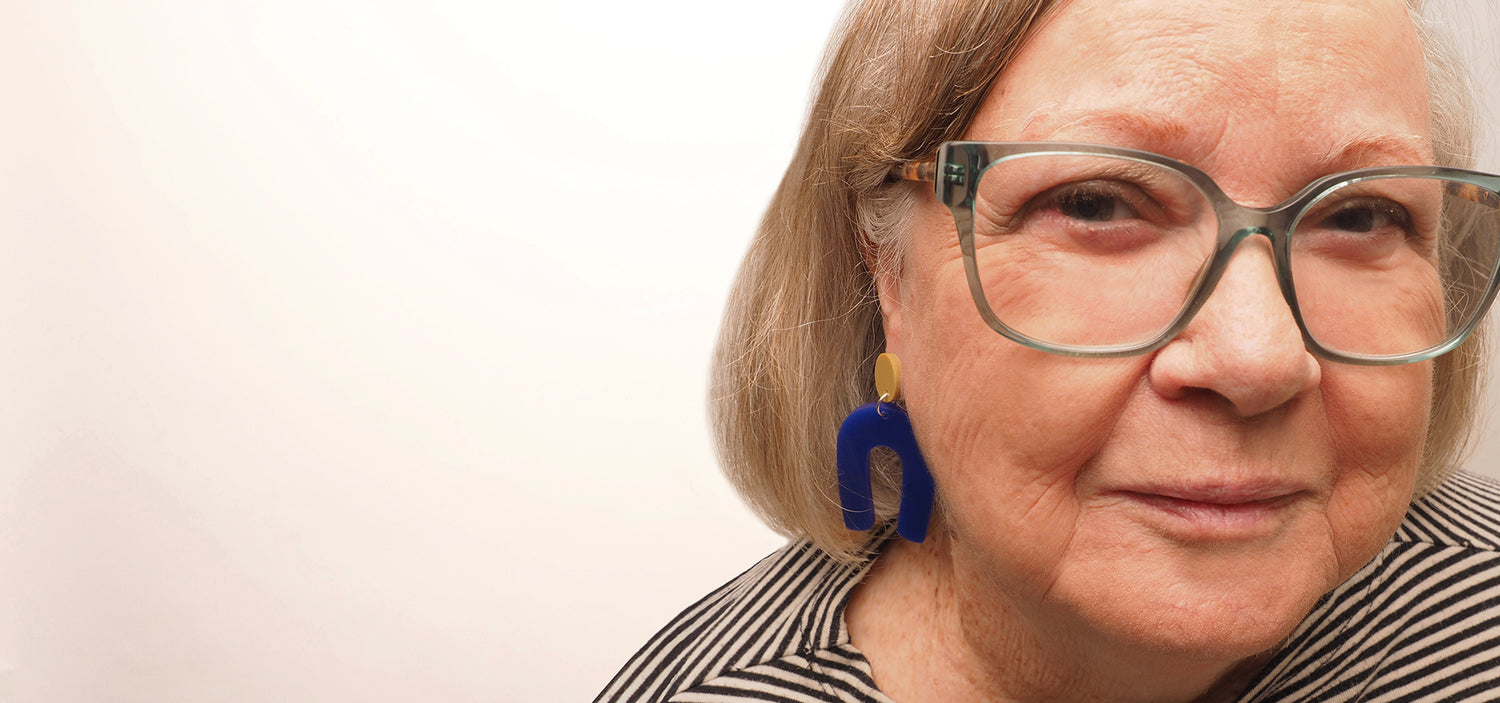 An older woman wearing navy blue arch earrings. The earrings are made with an olive coloured acrylic button, a navy blue acrylic arch, and silver surgical stainless steel jump rings.