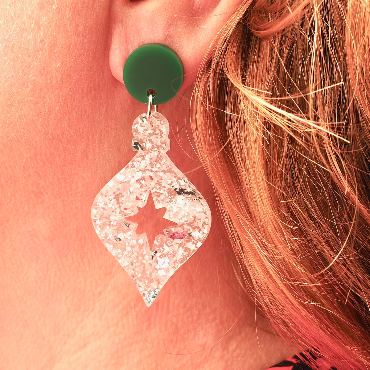 A close-up of a person's ear wearing a dangling earring. The earring has a round dark green top post and a clear, glittery teardrop-shaped drop with a star-shaped cutout in the center; this looks like a Christmas tree bauble.
