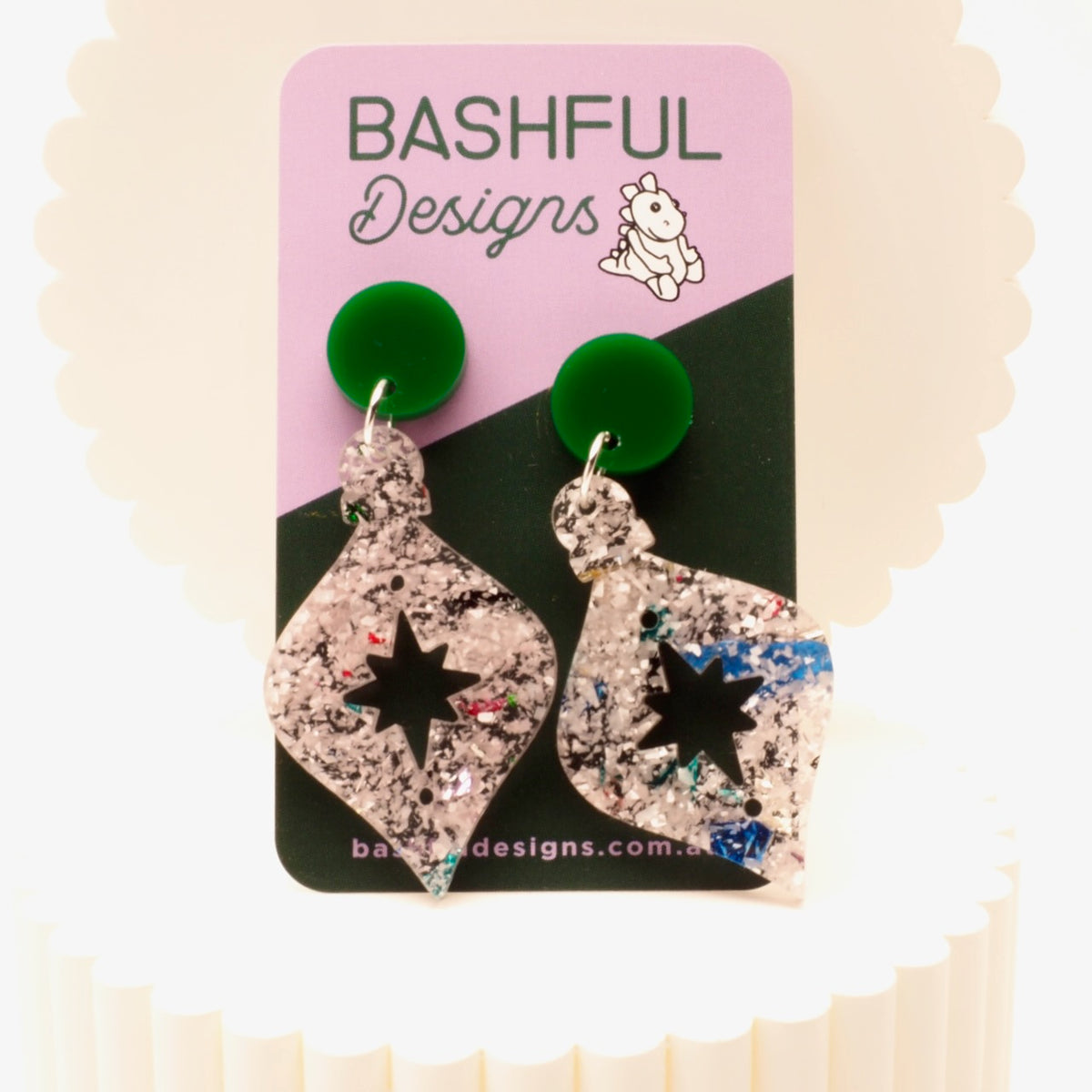 A pair of dangling earrings displayed on a pink and black card labeled Bashful Designs. The earrings have bright green round studs and teardrop-shaped charms covered in glittery silver, pink, and blue flecks, with a black star cutout in the center.