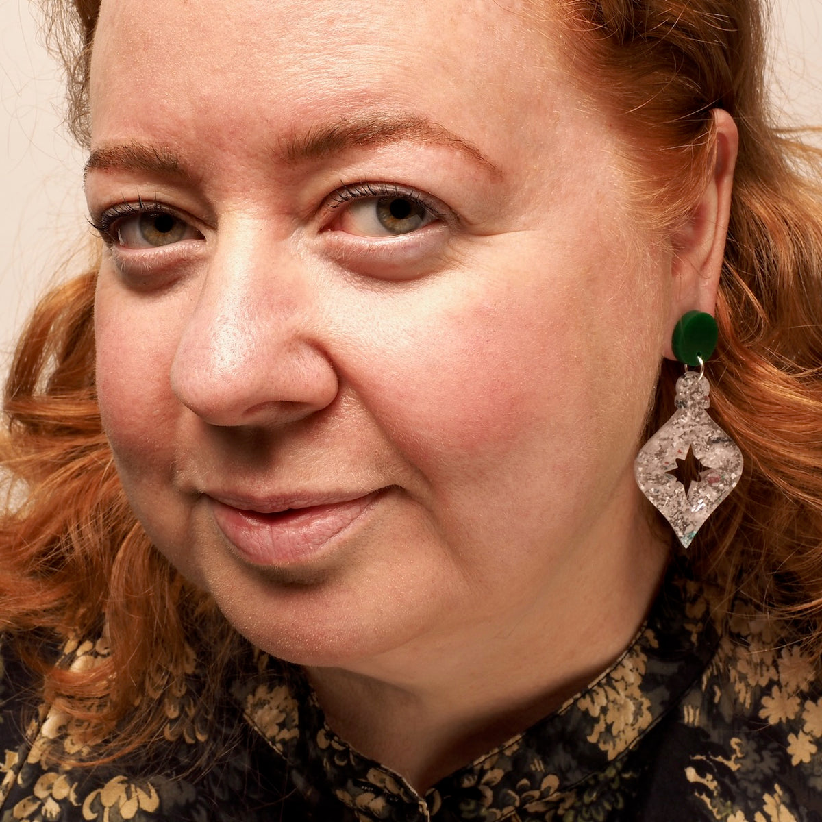 A close-up portrait of a woman with red hair wearing a distinctive dangling earring. The earring features a bright green circular top and a clear, diamond-like heart-shaped drop with a star cutout in the center; a Christmas bauble.