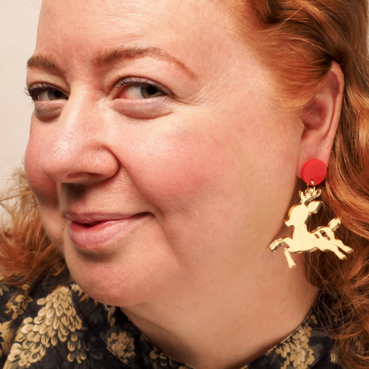 A close-up of a woman with red hair wearing a gold mirror  acrylic earring with a reindeer silhouette and a bright red circular top, against a plain light background.
