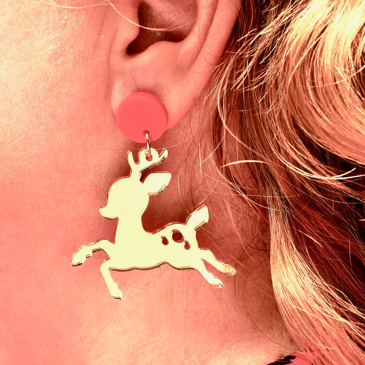 A close-up of a woman's ear, wearing a gold mirror acrylic reindeer earring, that has a red round stud top.