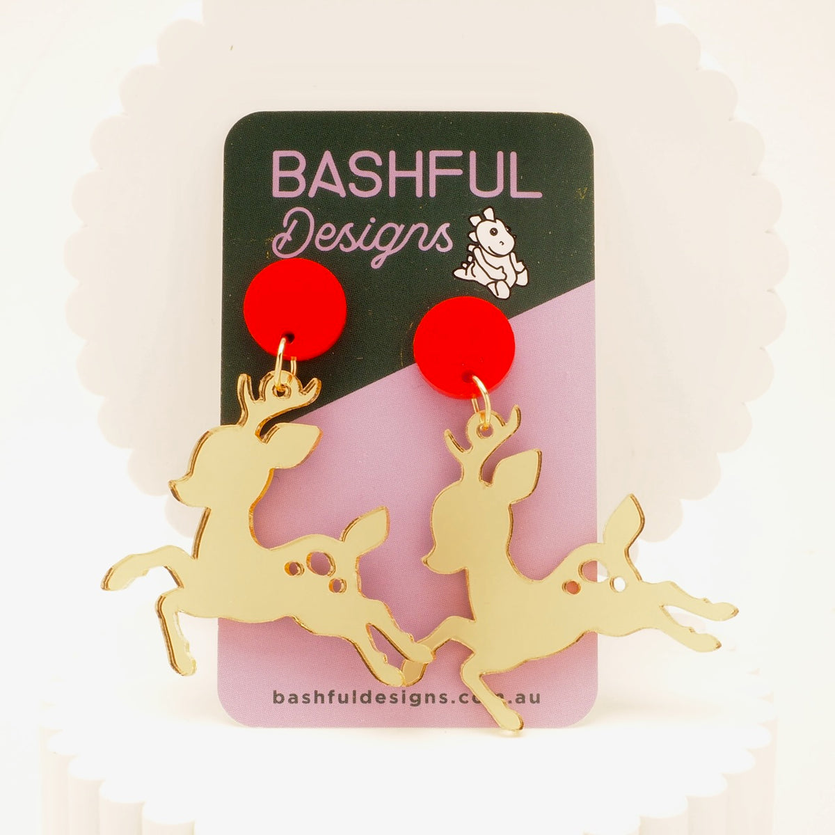 A pair of gold mirror acrylic earrings in the shape of Christmas reindeers, topped with red, round stud toppers.  These earrings are shown hanging from a Bashful Designs product card, and have a pale background.