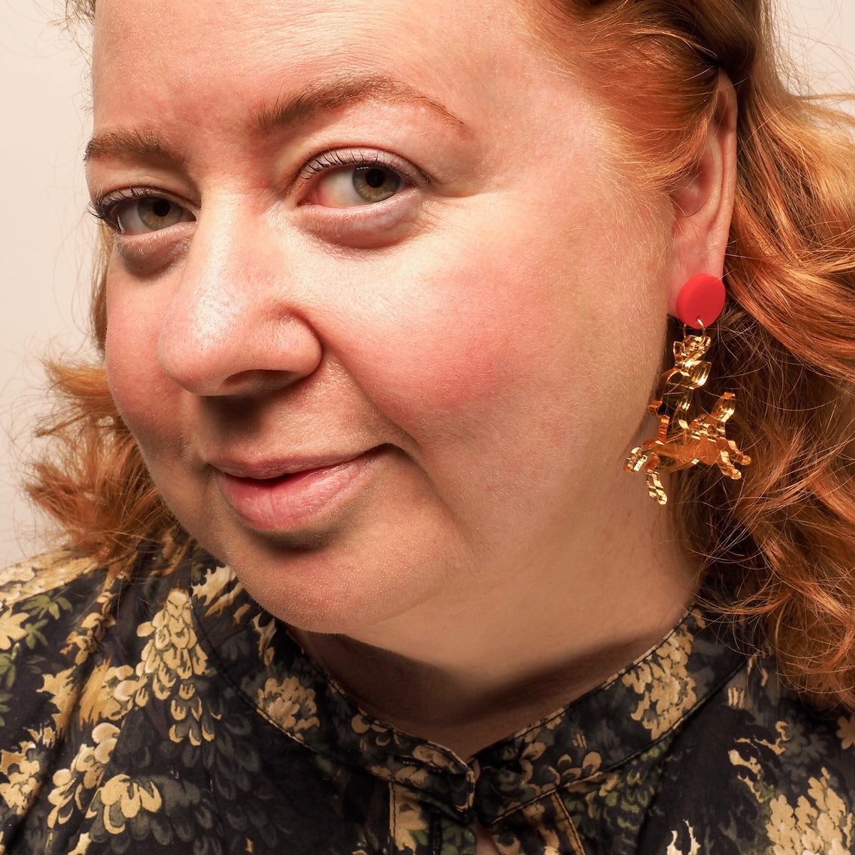A close-up portrait of a woman with red hair wearing a gold mirror acrylic dangling earring  in the shape of a Christmas reindeer, with a red circular top.