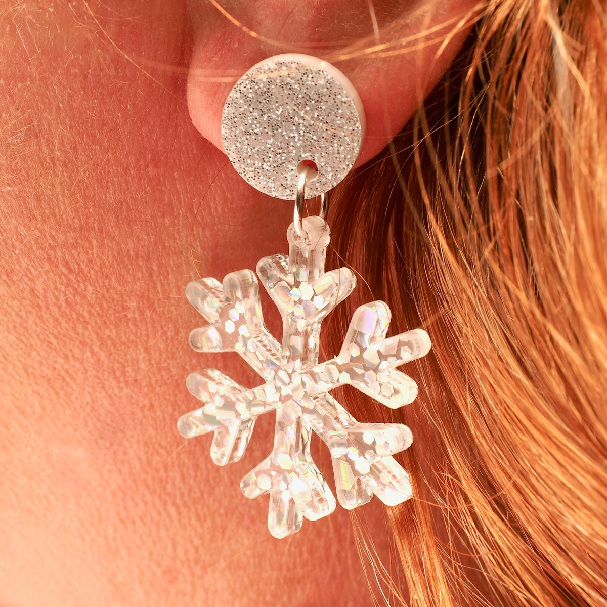 A close-up of a person's ear wearing a silver glitter round stud earring with a dangling clear snowflake charm filled with iridescent glitter.