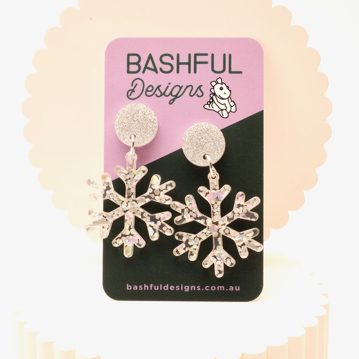 A pair of silver snowflake dangle earrings with clear rhinestones, displayed on a pink and black card from Bashful Designs.
