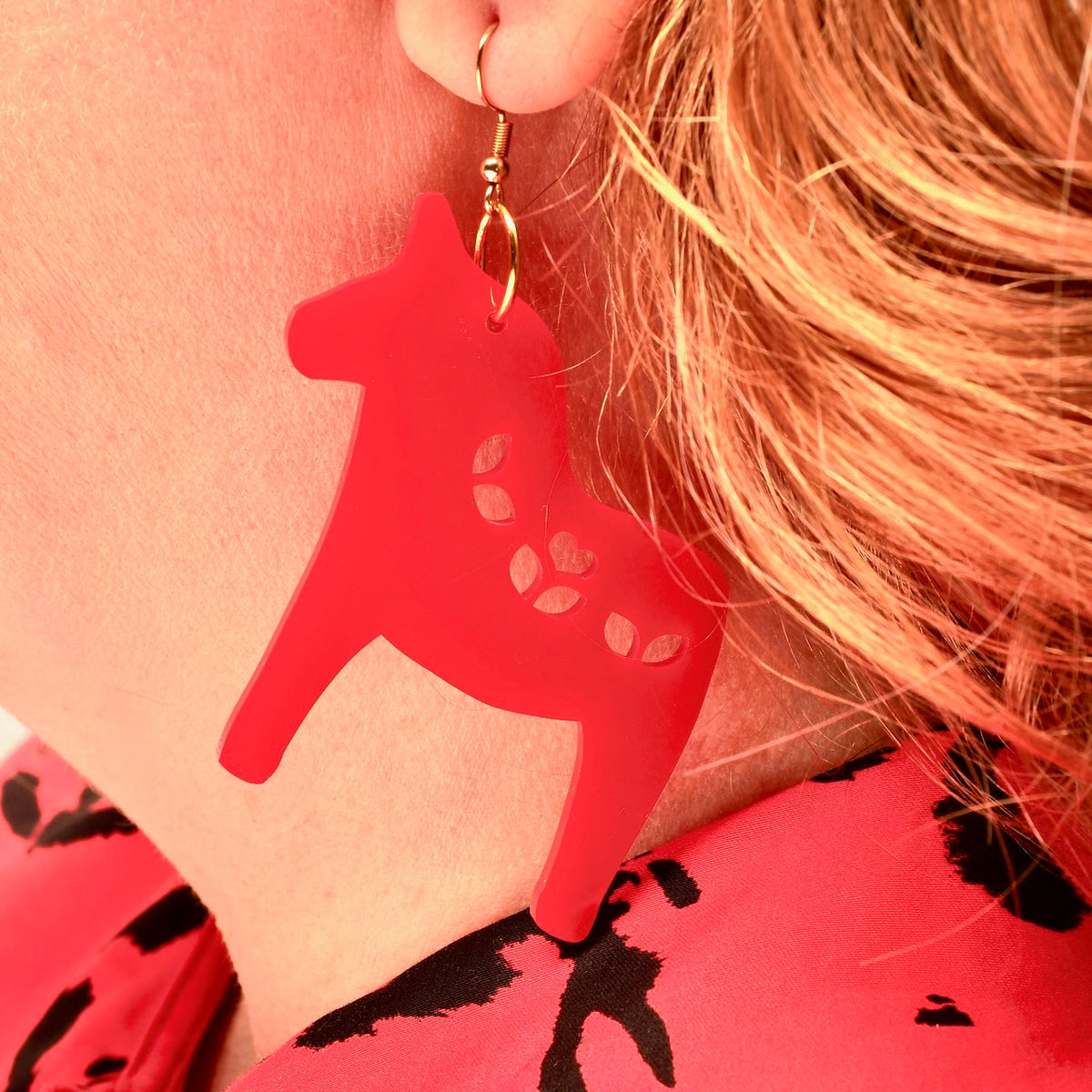 A close-up of a person wearing a bright red Dala horse-shaped acrylic drop earring with a gold fishhook wire.
