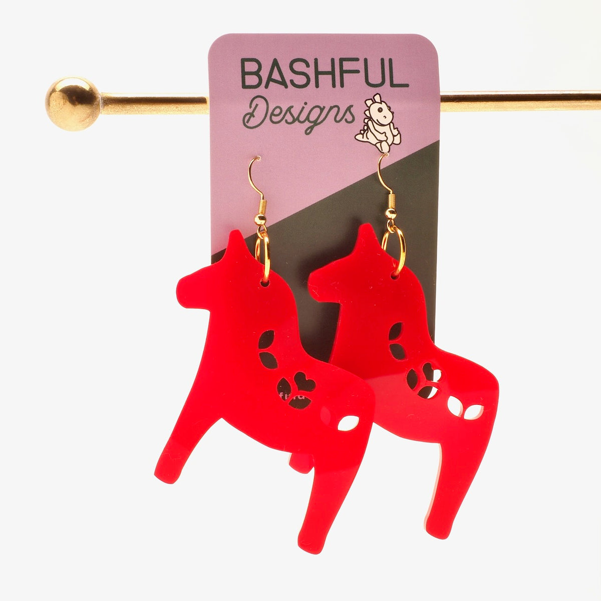 A pair of bright red Nordic Christmas Horse acrylic drop earrings with gold hooks, displayed on a pink and black 'Bashful Designs' backing card, hung from a gold display rod against a white background.