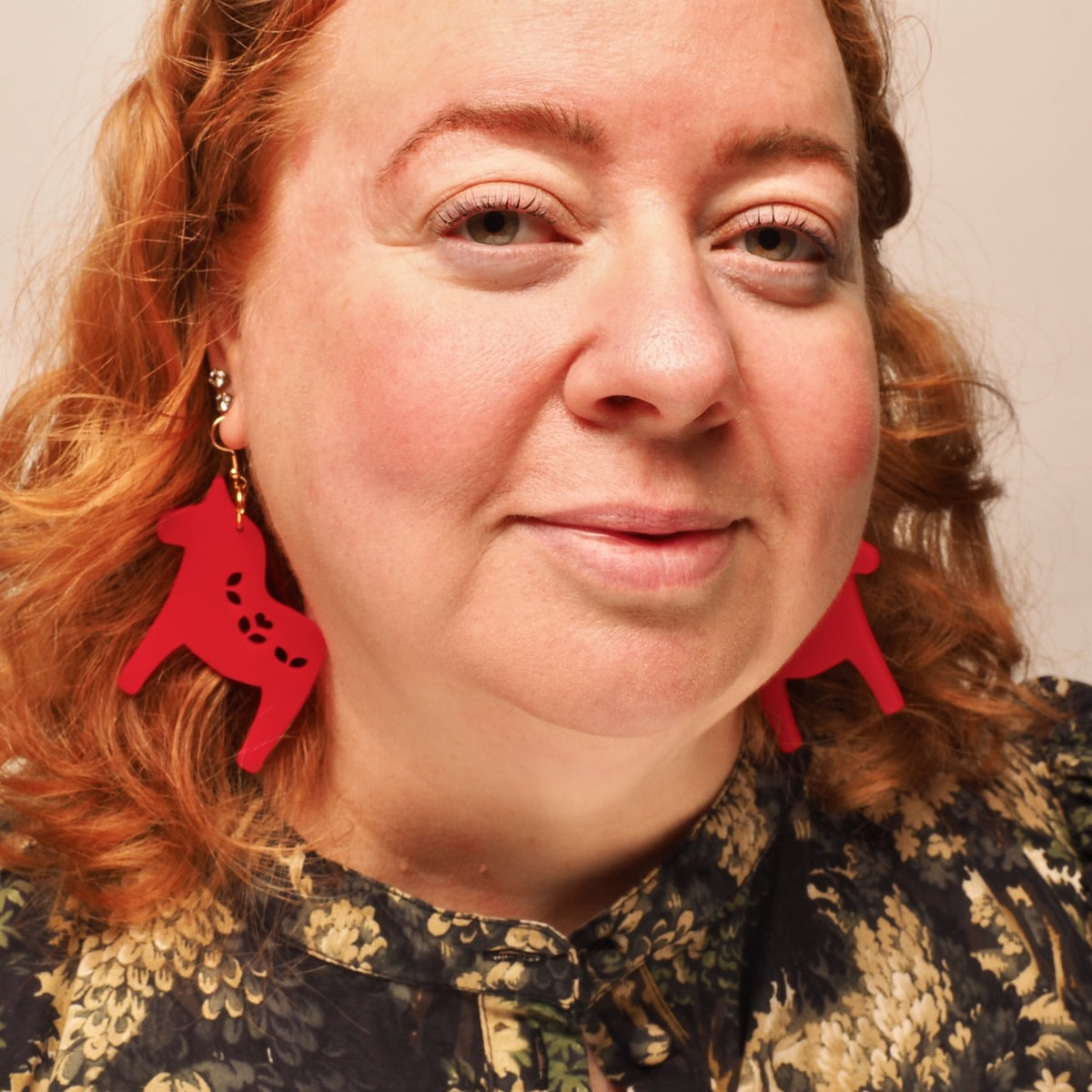 A close-up portrait of a woman with red hair wearing bright red Nordic Christmas horse-shaped earrings.
