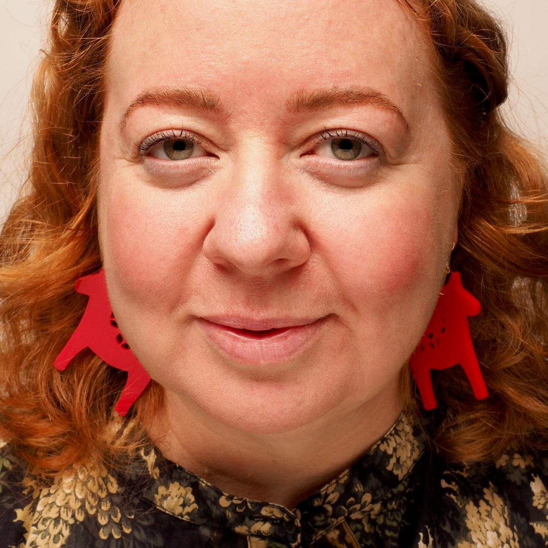 A close-up portrait of a woman with red hair wearing bright red acylic earrings, in the shape of a Nordic Christmas Horse.