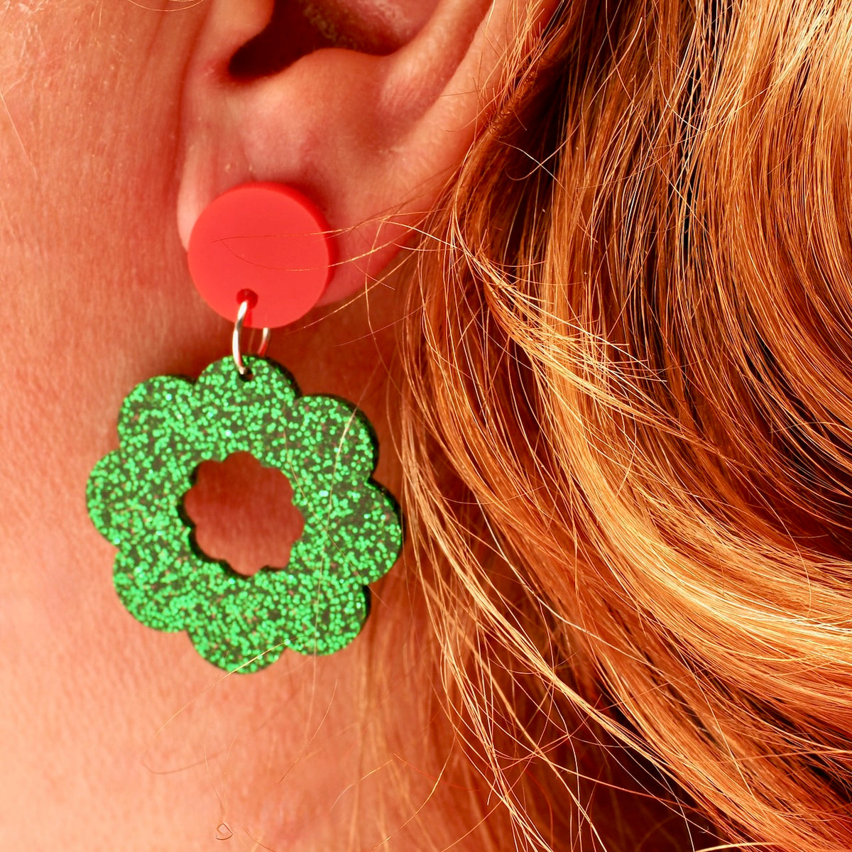 A close-up of a person's ear wearing a bright red circular stud earring with a dangling glittery green Christmas wreath shaped drop.