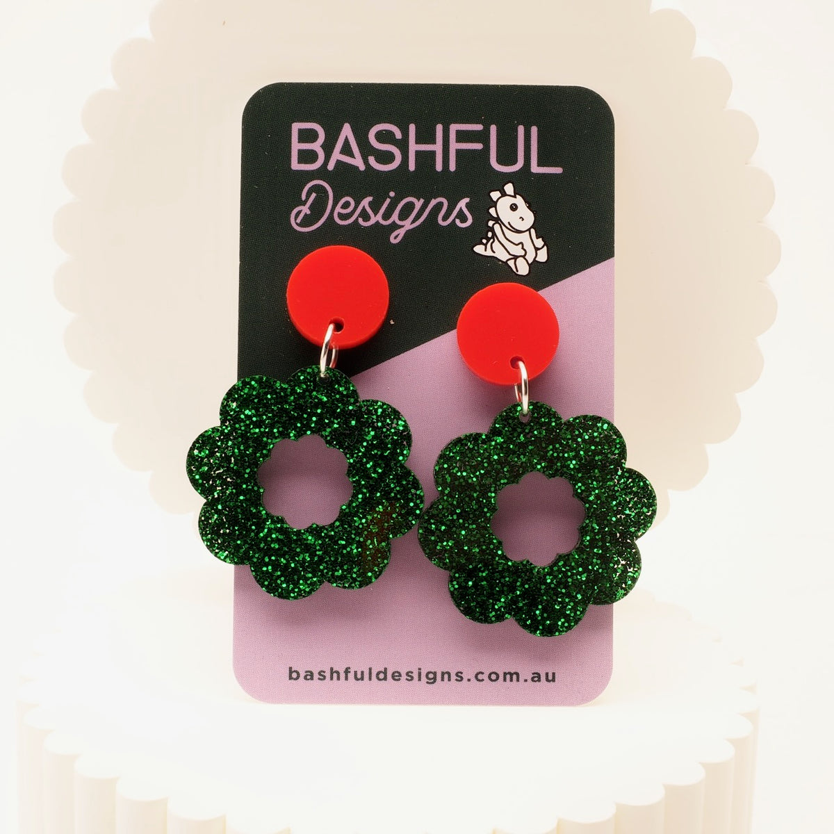 A pair of green glitter Christmas wreath shaped drop earrings with bright red studs, displayed on a pink and black card labeled 'Bashful Designs'.
