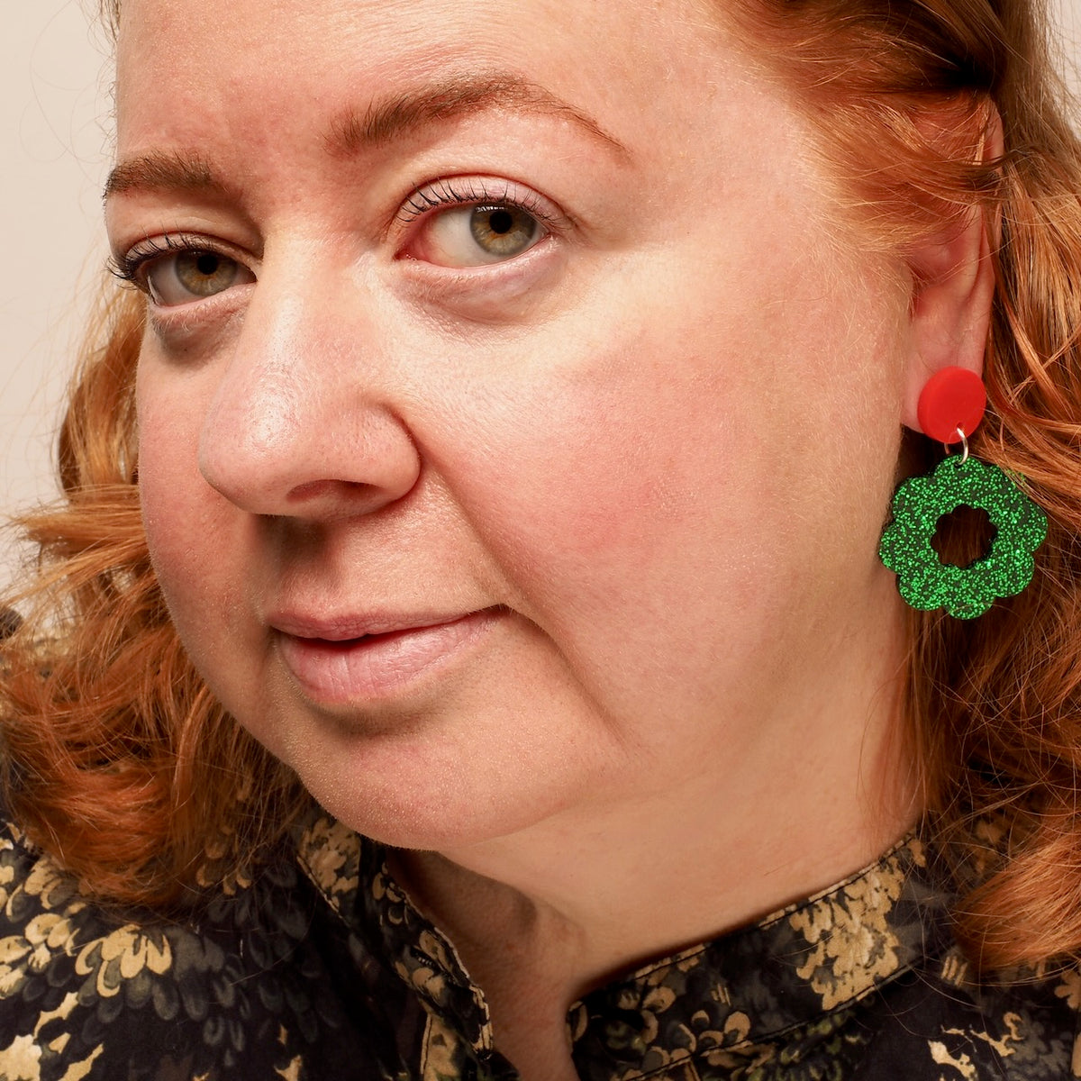 A close-up portrait of a woman with red hair wearing a green glittery Christmas wreath shaped earring with a red top disc, against a light neutral background.