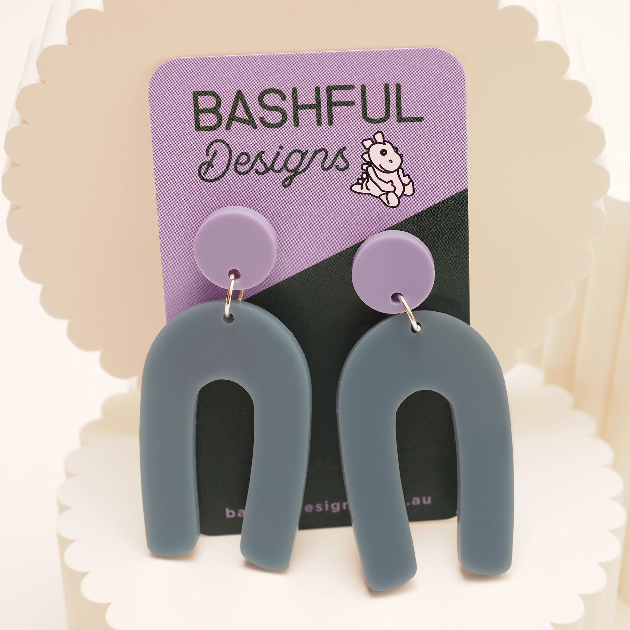 Grey-ish blue arch shaped earrings. They feature a round, lilac earring topper and are shown on a 'Bashful Designs' earring card.