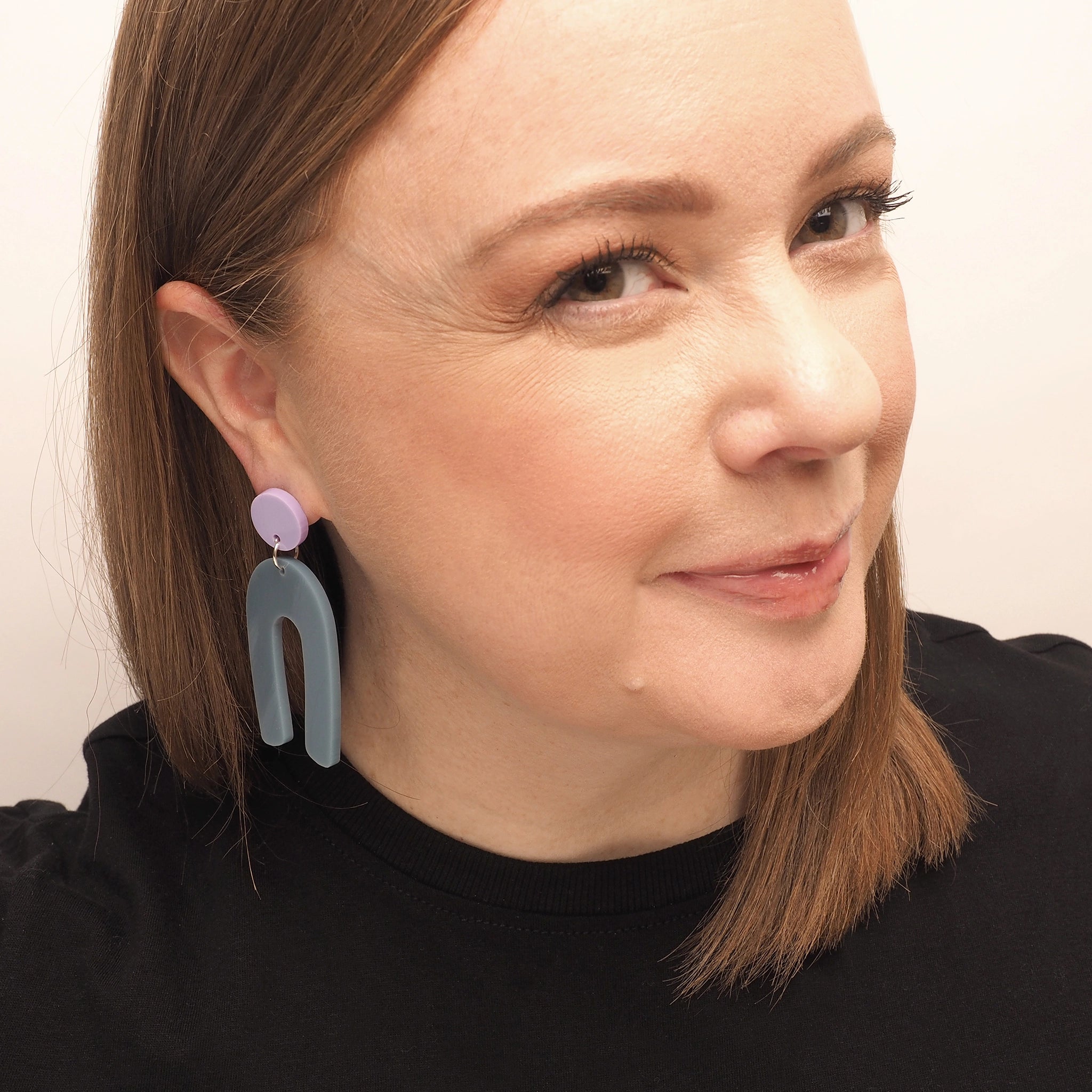Grey-ish blue arch shaped earrings with a round, lilac earring topper shown on a curvy woman with straight red hair.