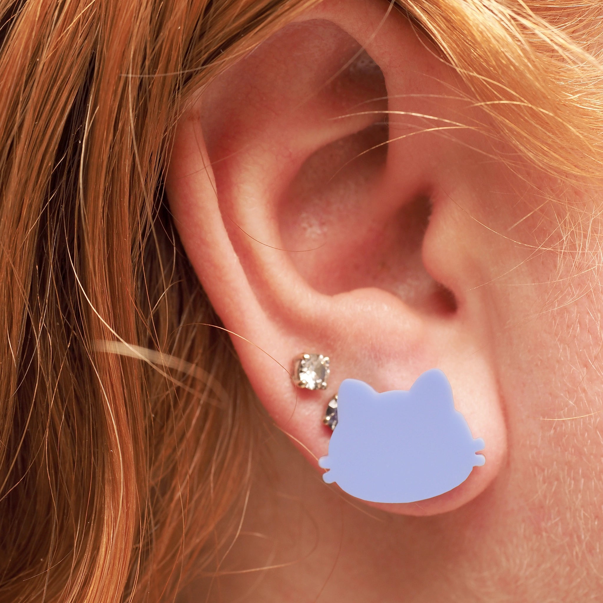 A woman with curly red hair wearing light blue stud earrings in the shape of a cat.