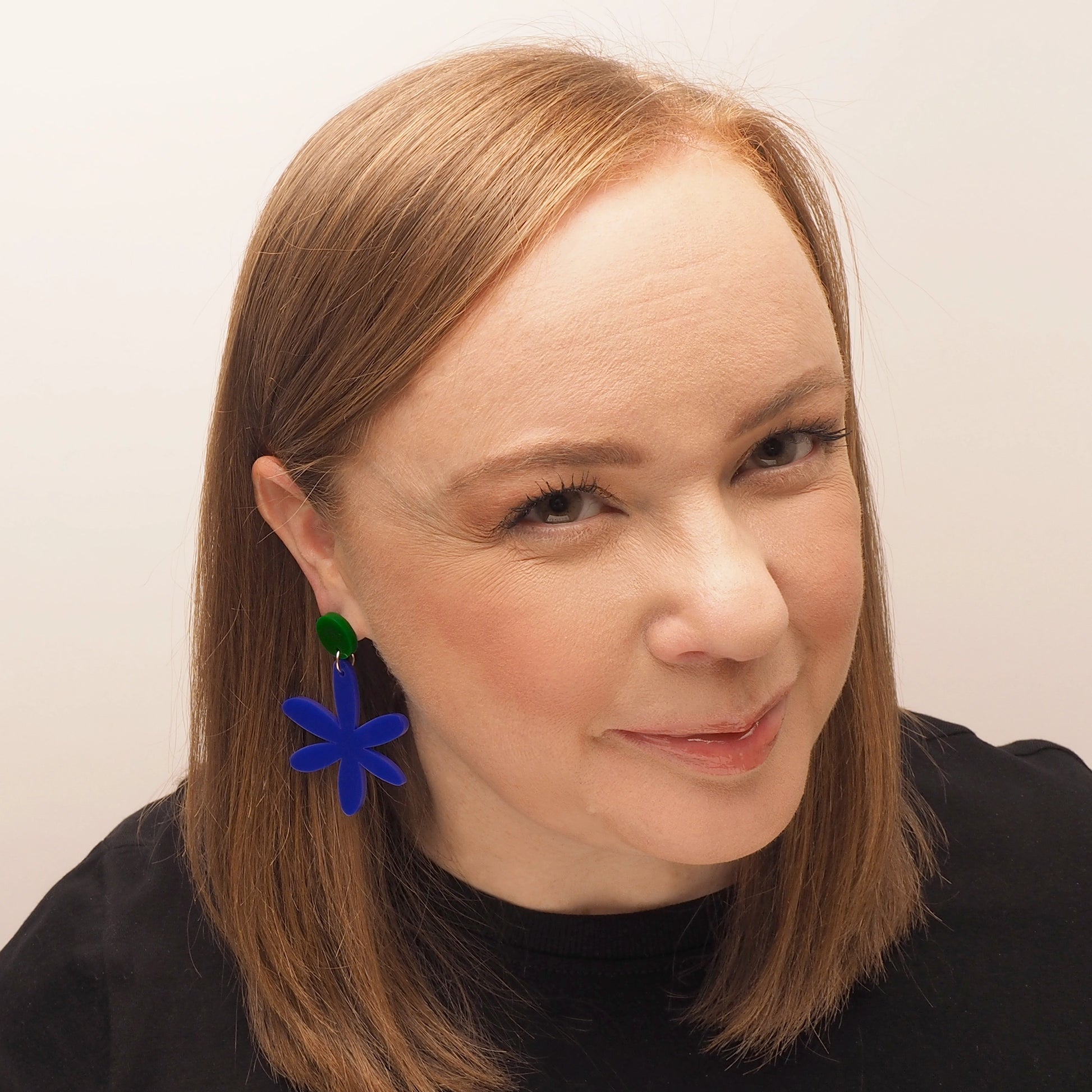 A curvy woman with red hair wearing navy blue acrylic earrings in the shape of a flower. These earrings are topped with a dark green round.