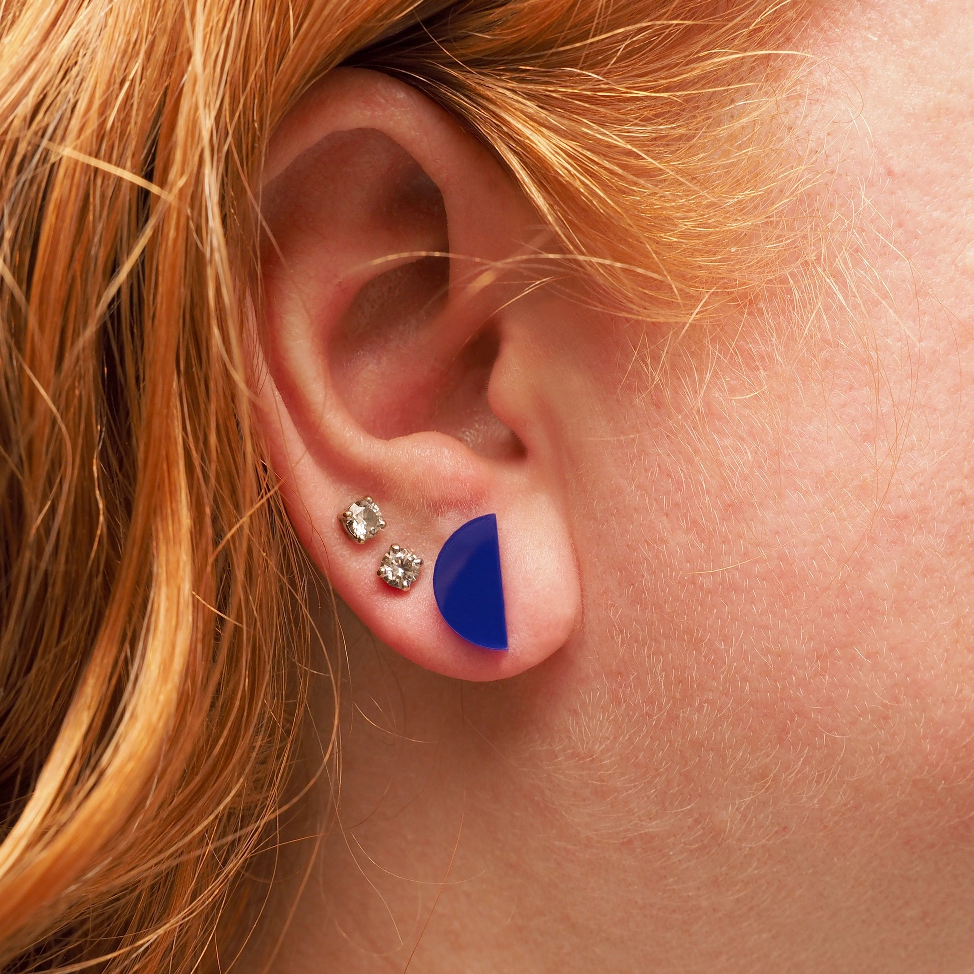A person with curly red hair wearing navy acrylic stud earrings in the shape of a half-moon.