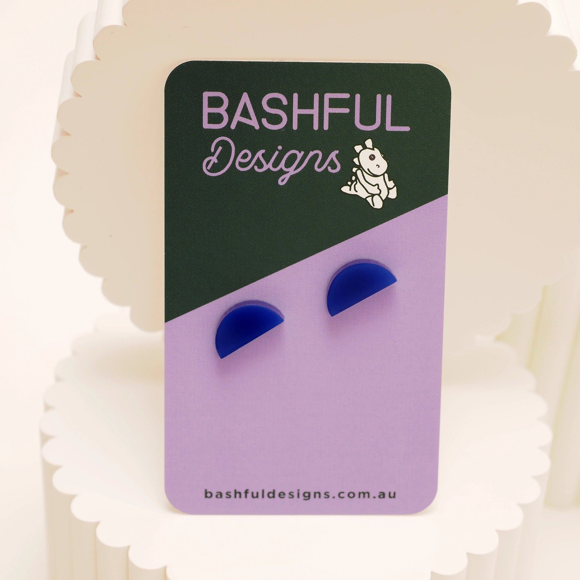 A pair of navy acrylic stud earrings in the shape of a half-moon. The earrings are shown on a 'Bashful Designs' branded card.