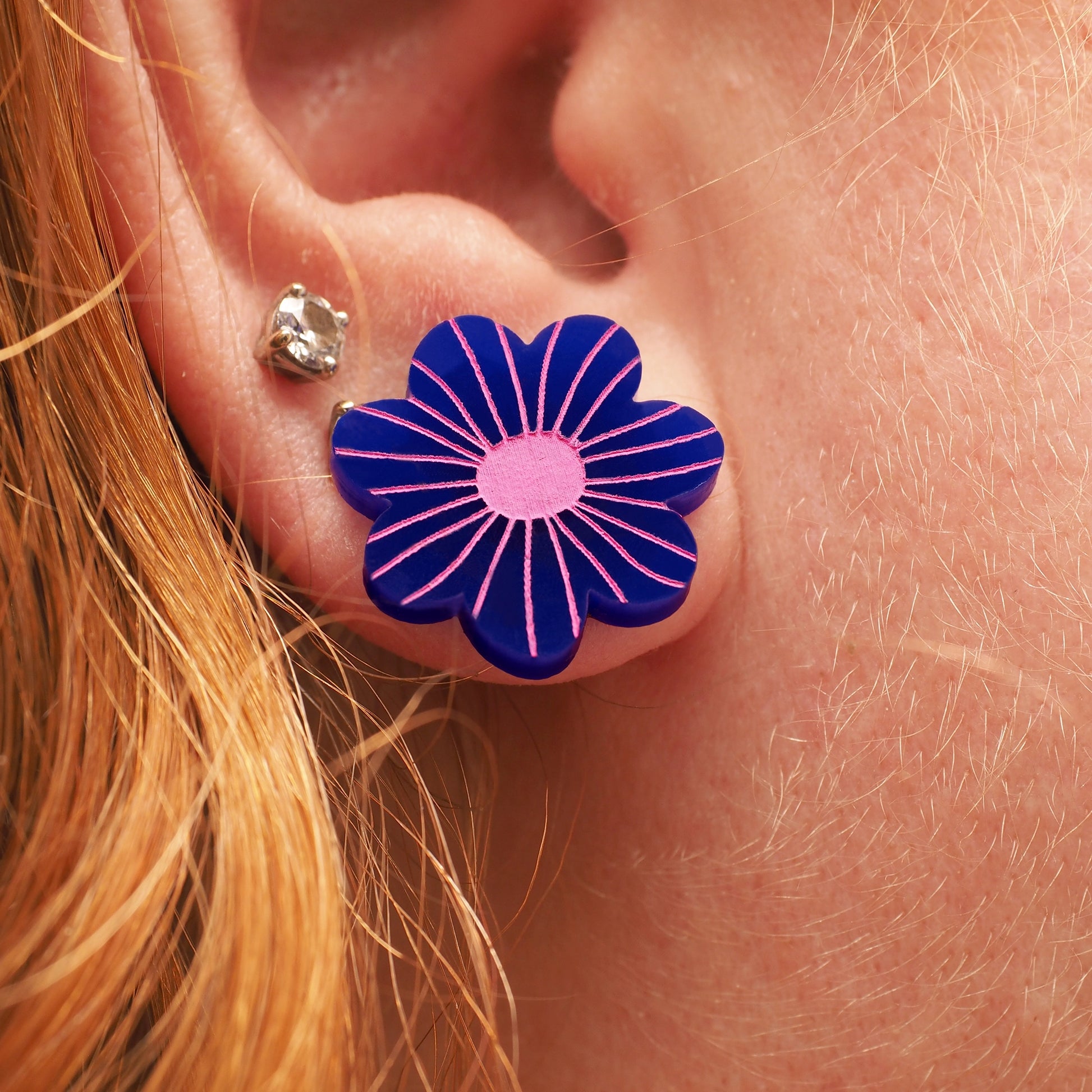 A person with red and blonde hair wearing a navy acrylic flower shaped stud earring, that has pink engraving.