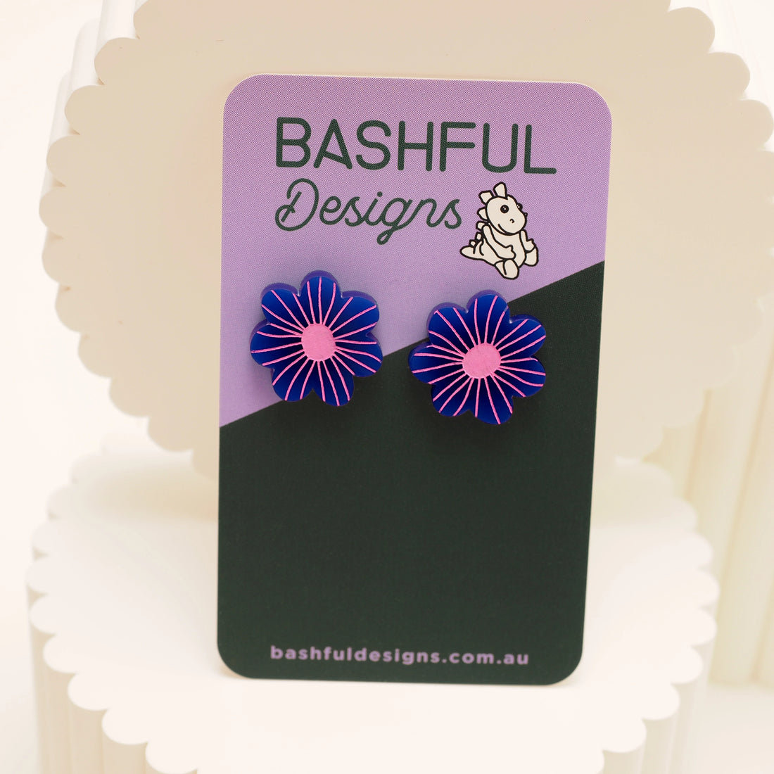 Flower-shaped stud earrings on a 'Bashful Designs' branded card with a pink background. These studs are navy blue, engraved with a light pink.