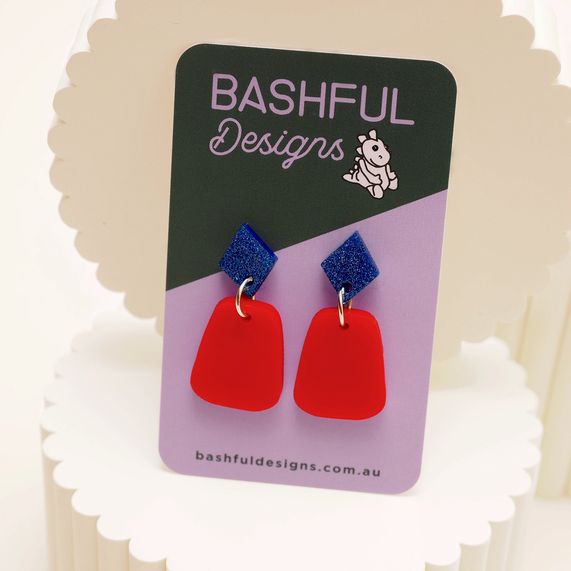 A pair of small dangles featuring blue and red acrylic. The topper is blue glitter acrylic in a diamond shape. Sitting below the blue diamond is a solid red acrylic oblong shape.