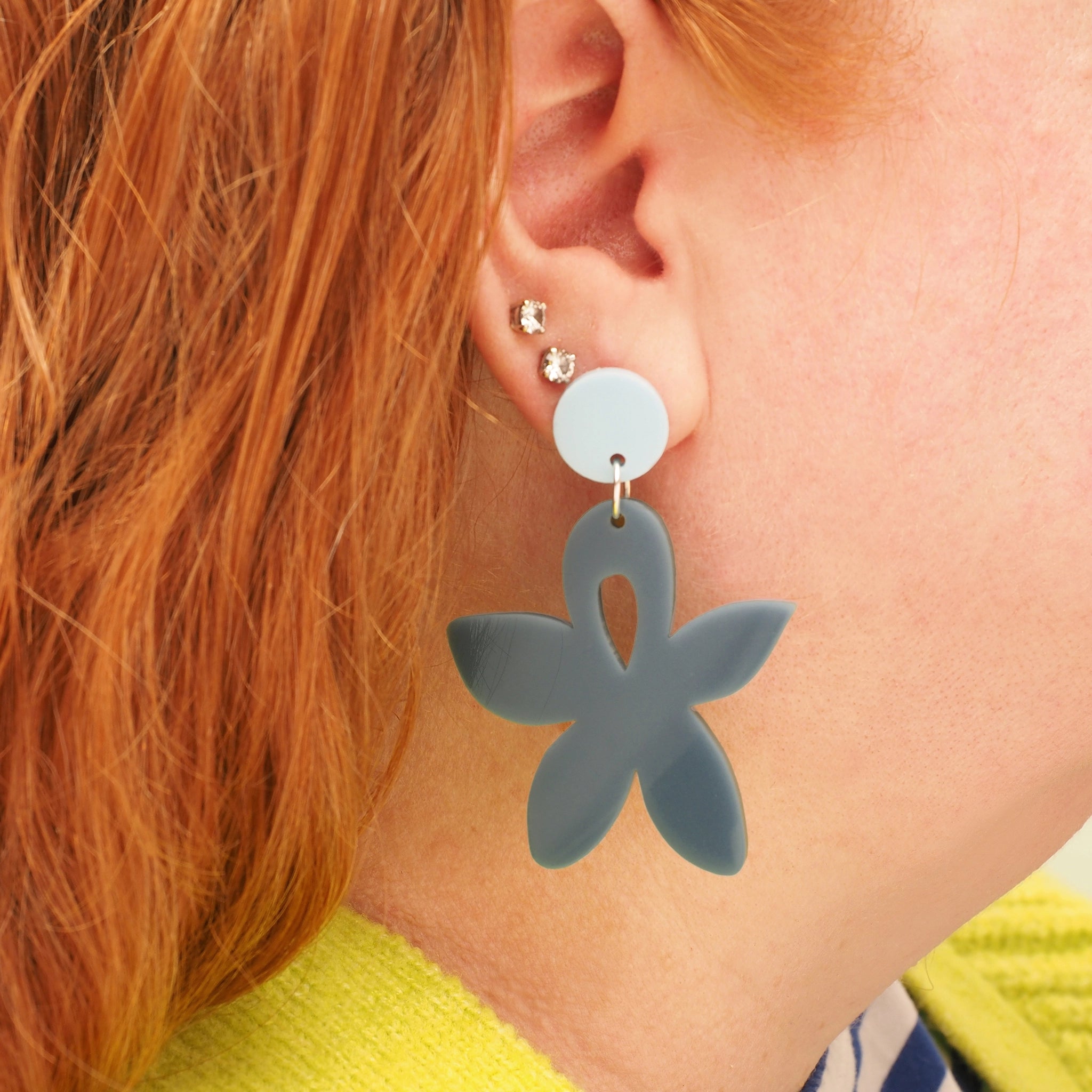 A person with long red hair wearing a wildflower shaped earring that is grey-ish blue with a light blue, round earring topper.