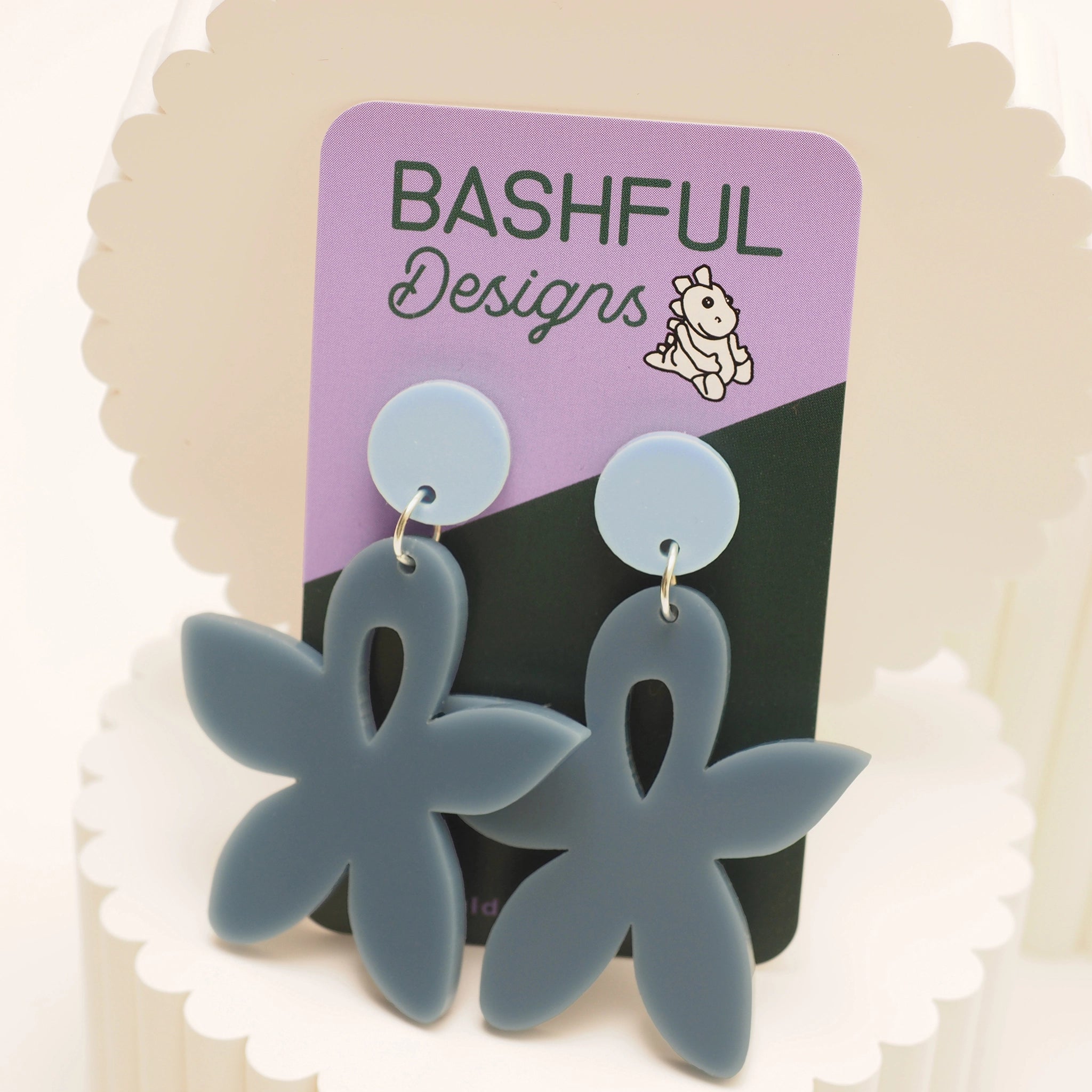 A pair of grey-ish blue acrylic earrings shaped like a wild flower and with a light blue, round earring topper. The earrings are shown on a 'Bashful Designs' branded card.