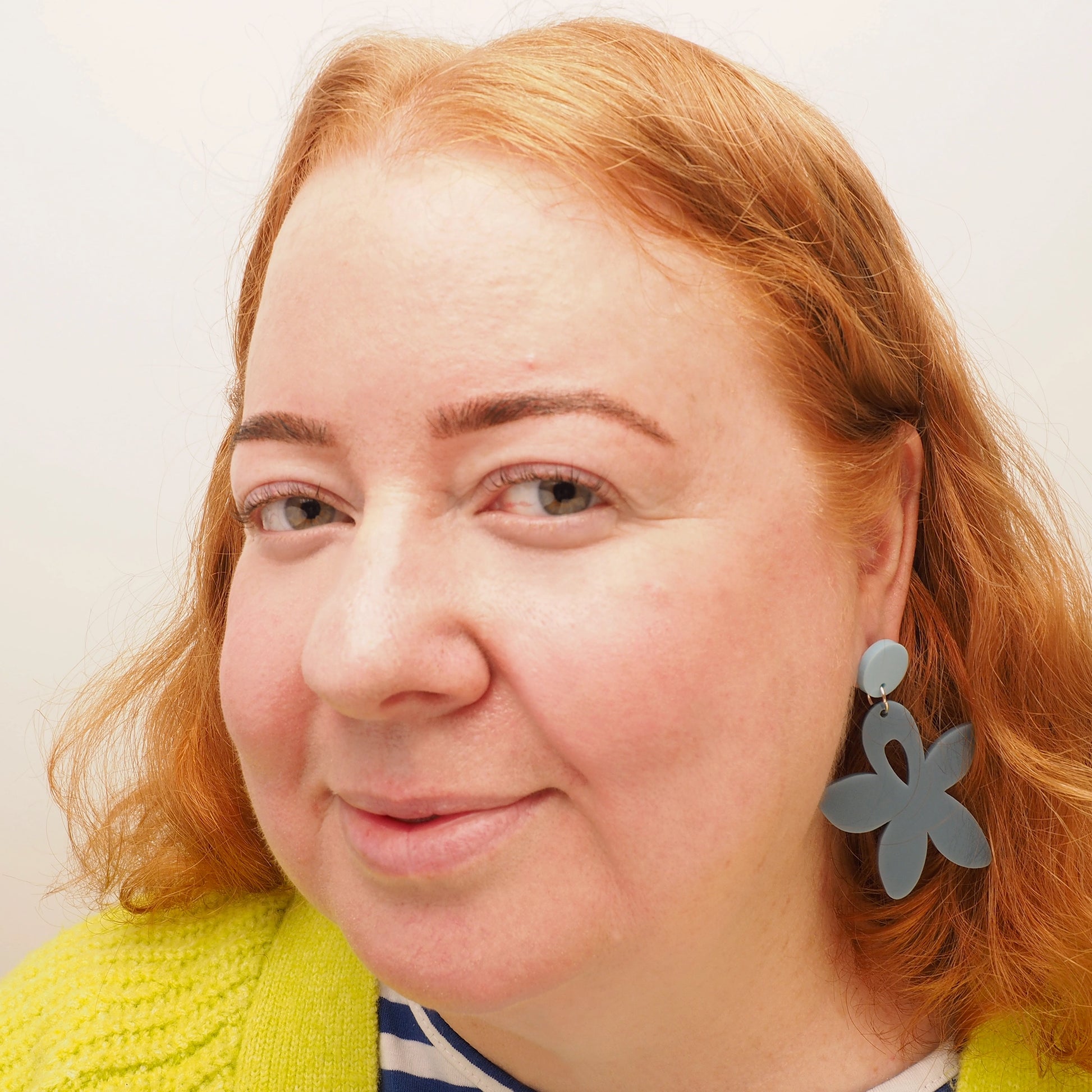 A curvy woman with long red hair wearing grey-ish blue wildflower shaped earrings with a light blue, round earring topper.