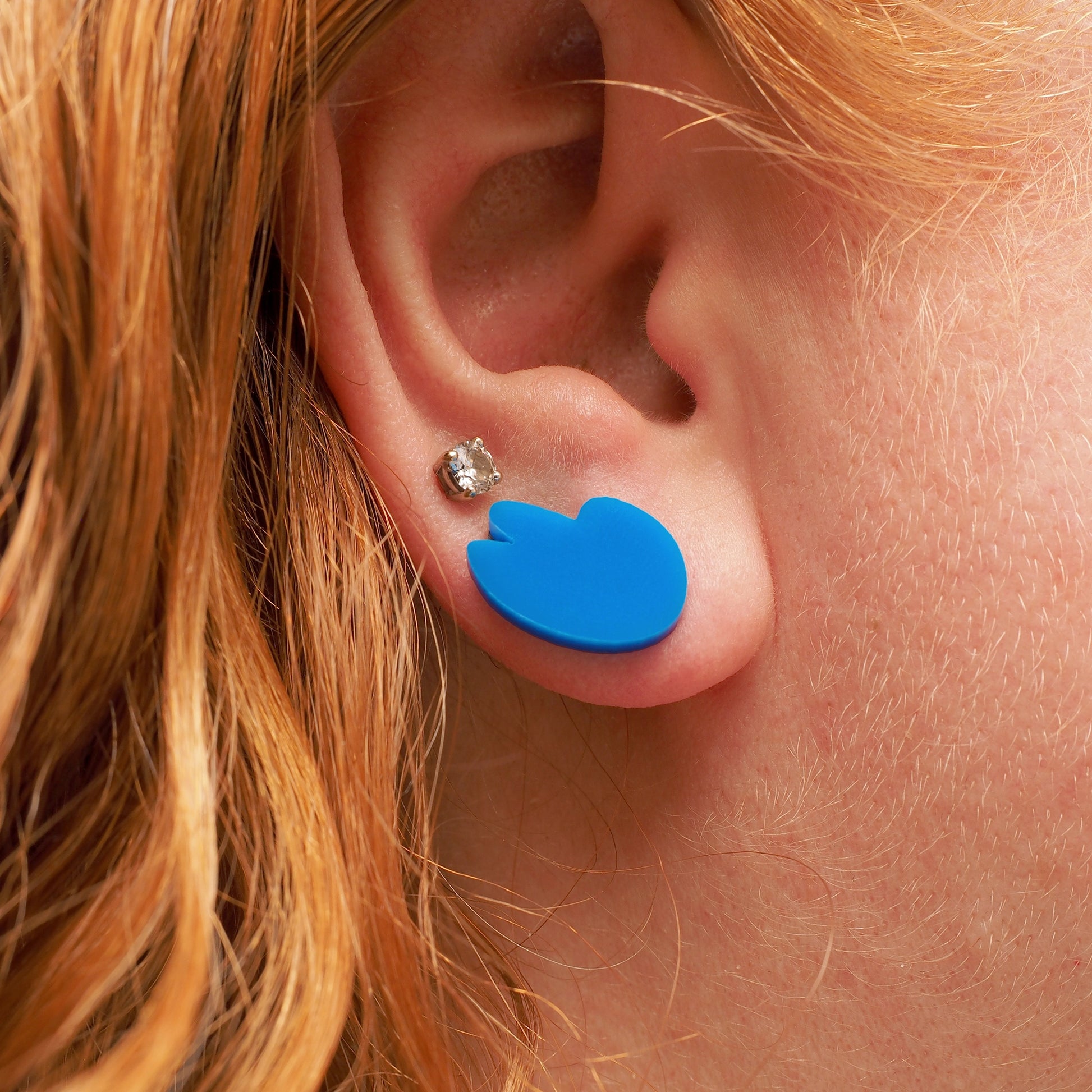 A person with curly red hair wearing bright blue acrylic stud earrings in the shape of a bird's wing.