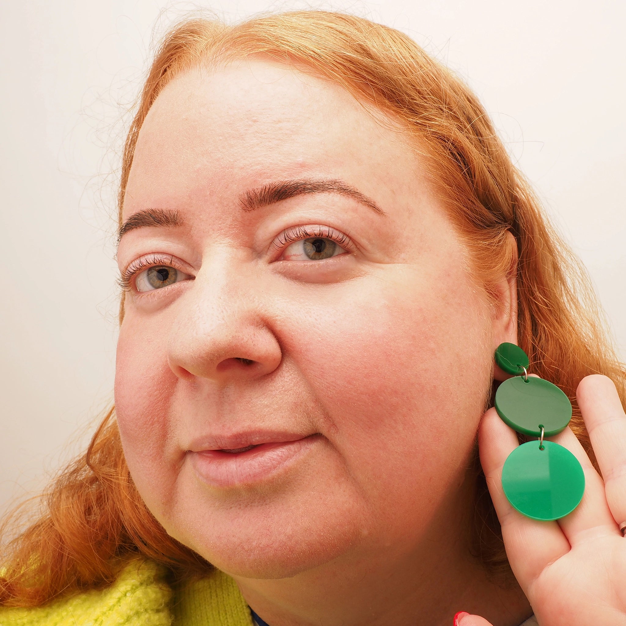 A woman with red hair wearing a pair of green chandelier earrings. The earrings consist of three circles, one small, followed by two medium circles. The top two circles are dark green and the bottom circle is a mid-to-light green.