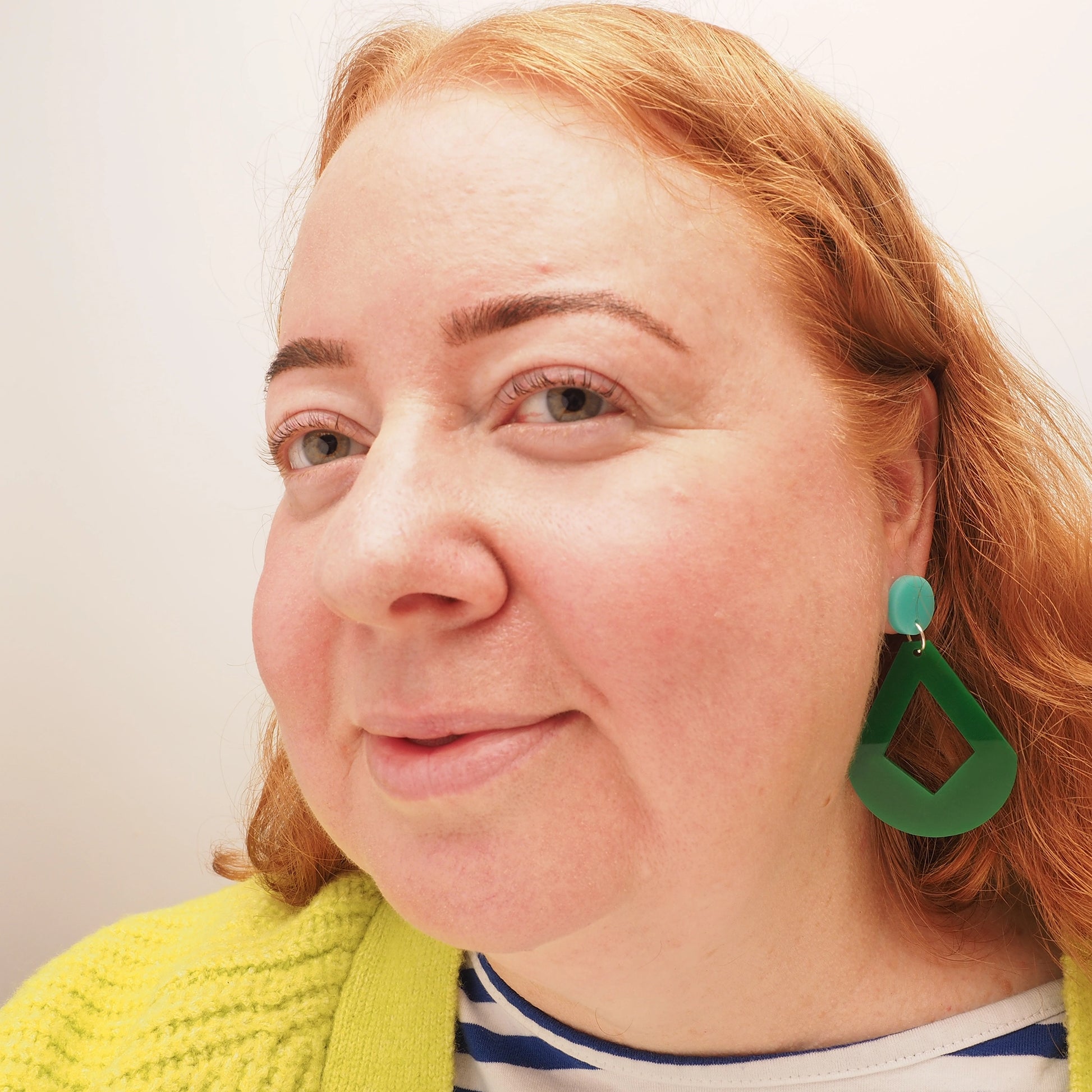 A curvy woman with curly red hair wearing green acrylic dangle earrings in the shape of a curvy diamond. These earrings feature an aqua earring topper.