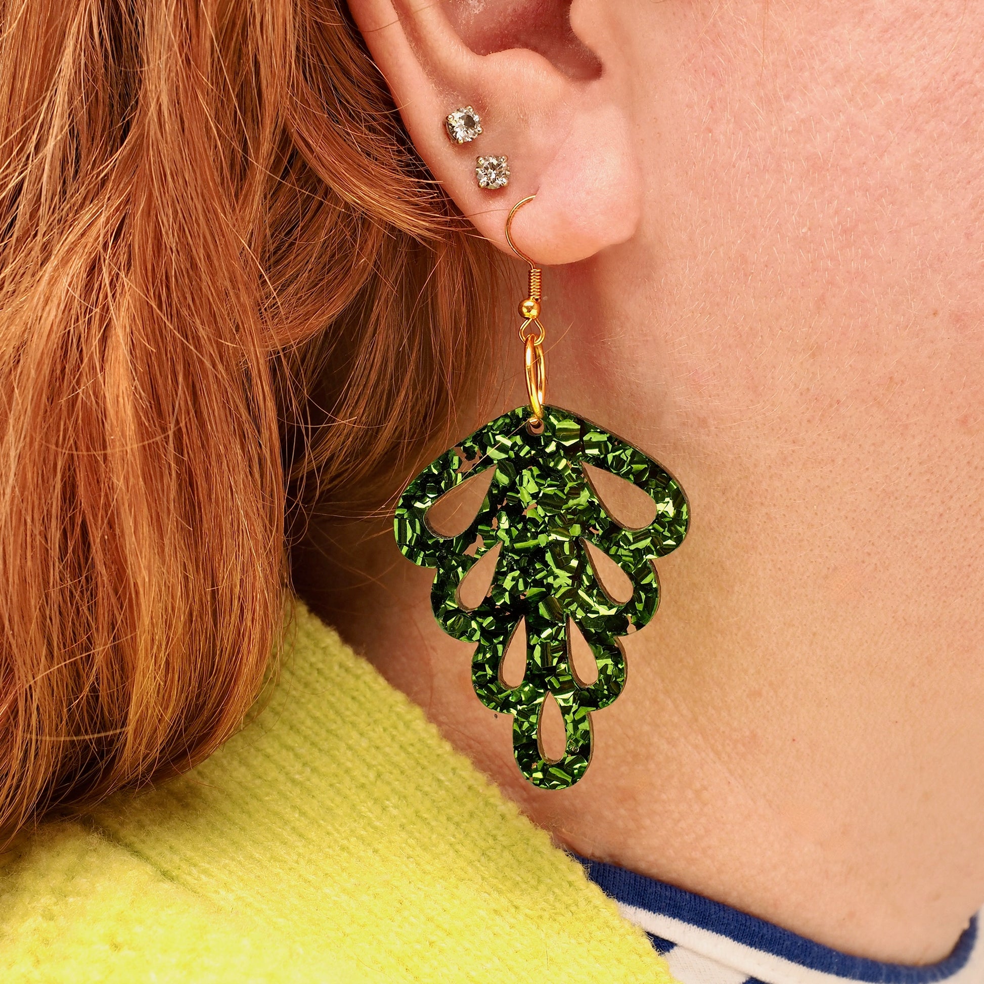 A woman with long red hair wearing green glitter acrylic earrings in the shape of a leaf.