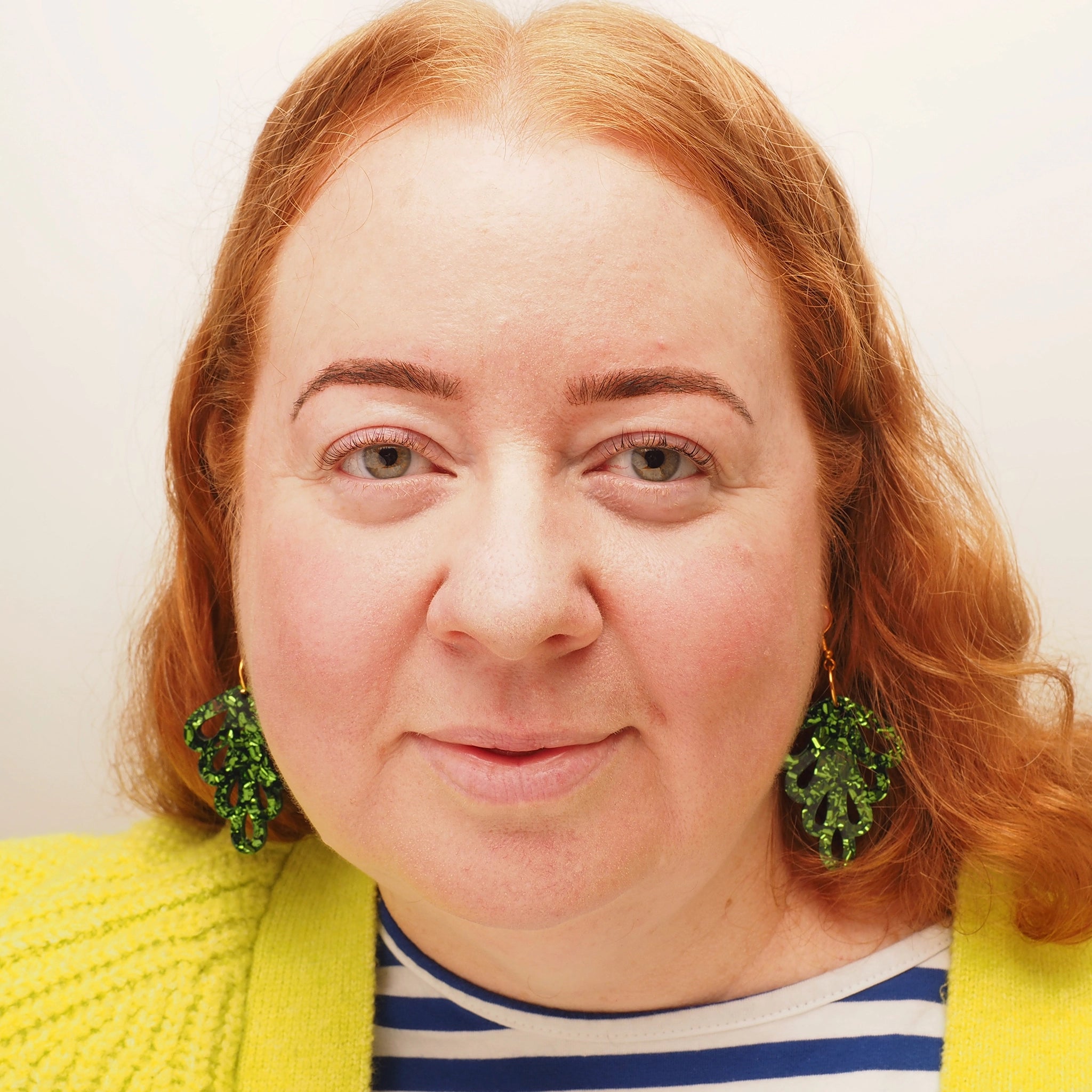 A curvy woman with long red curly hair wearing a pair of green glitter earrings in the shape of a leaf.