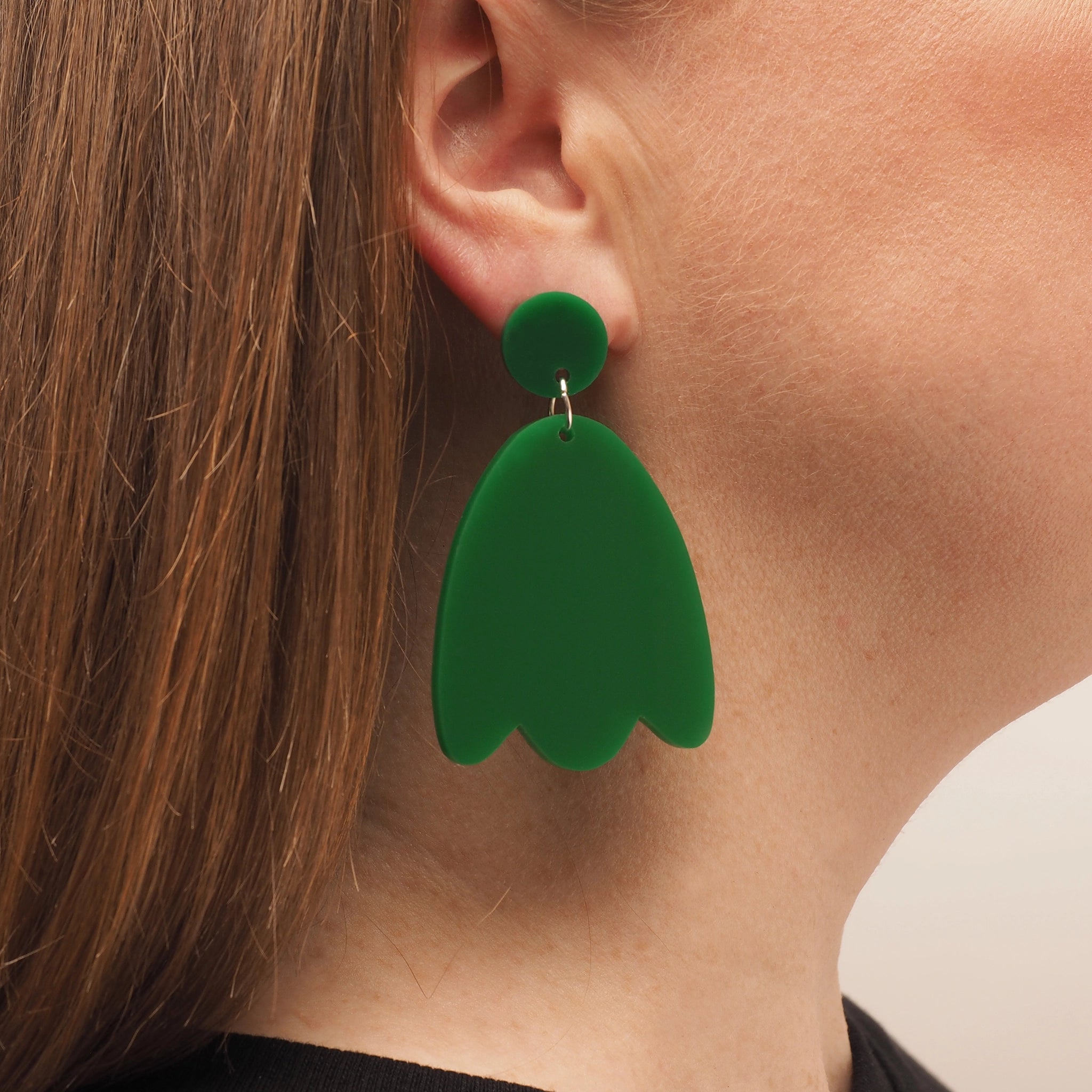 A person with straight red hair wearing a solid green acrylic earring in the shape of a petal.