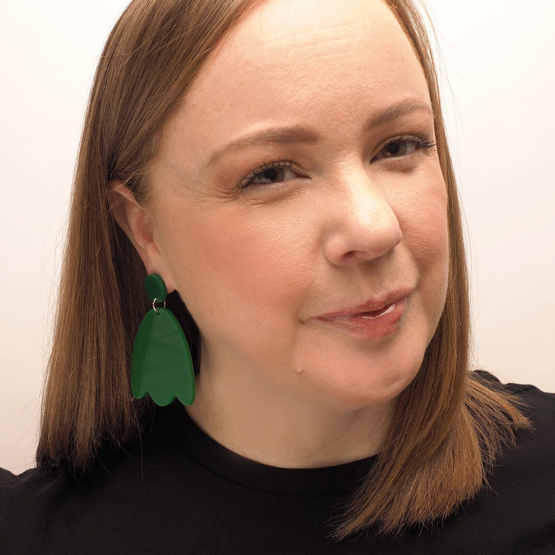 A woman with long straight red hair wearing a pair of solid green acrylic earrings in the shape of a petal.