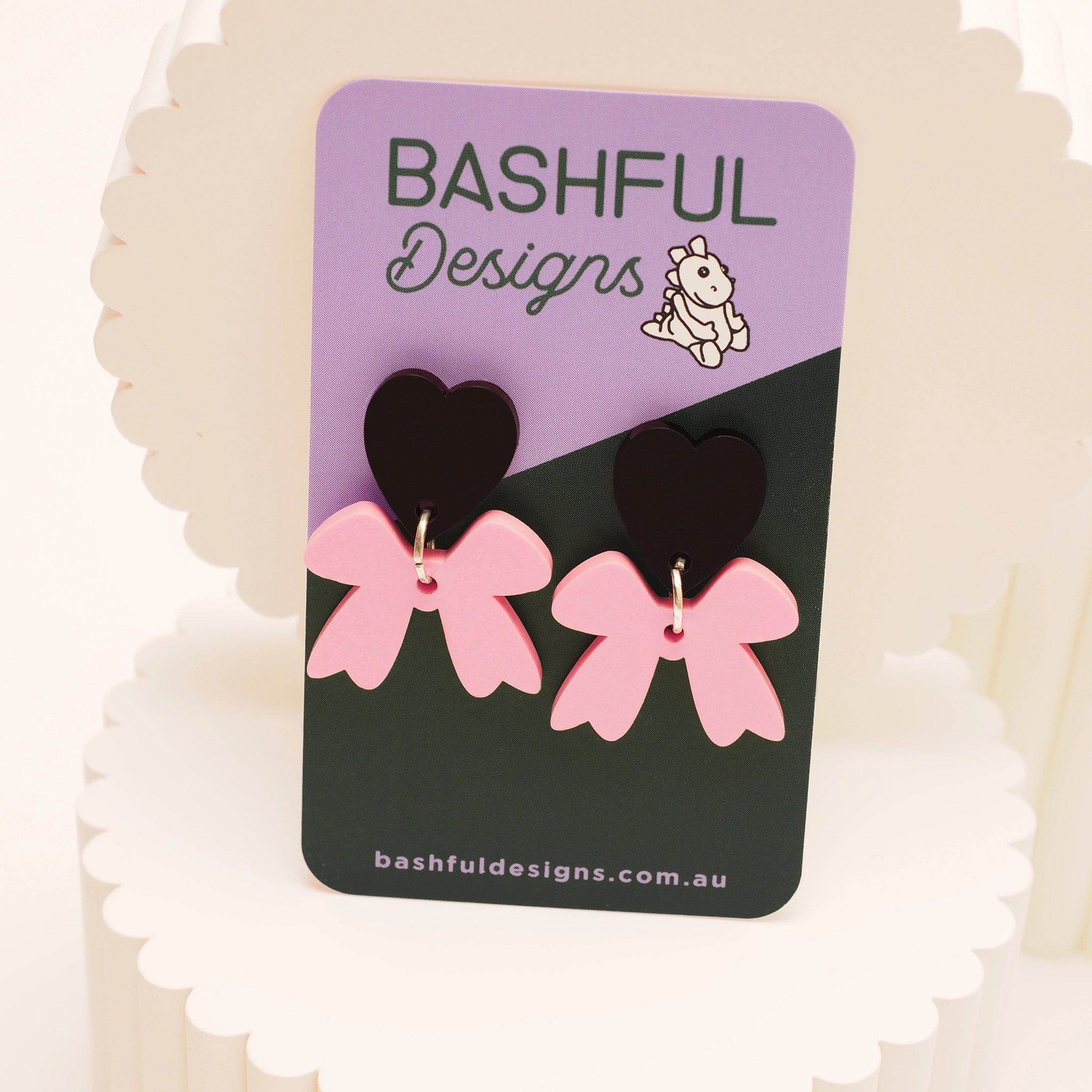 Black heart-shaped acrylic stud dangle earring, with a pink bow underneath the heart, shown on a 'Bashful Designs' product card.