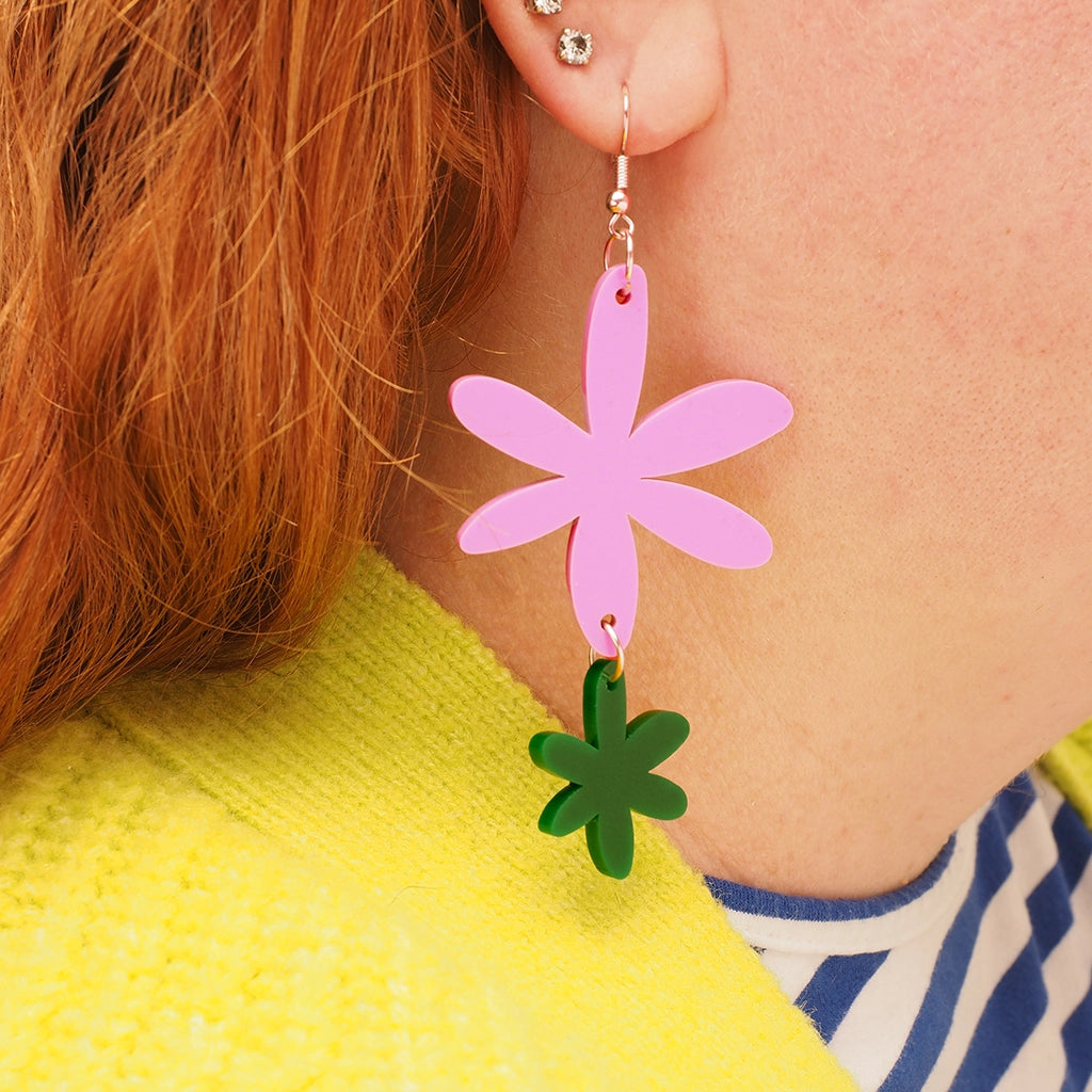 A close-up of a red-headed model wearing the pink and green floral earrings in her ear. it is a side-on, profile view of the earrings, which are made of a large pink acrylic daisy and a small, dark green acrylic daisy.