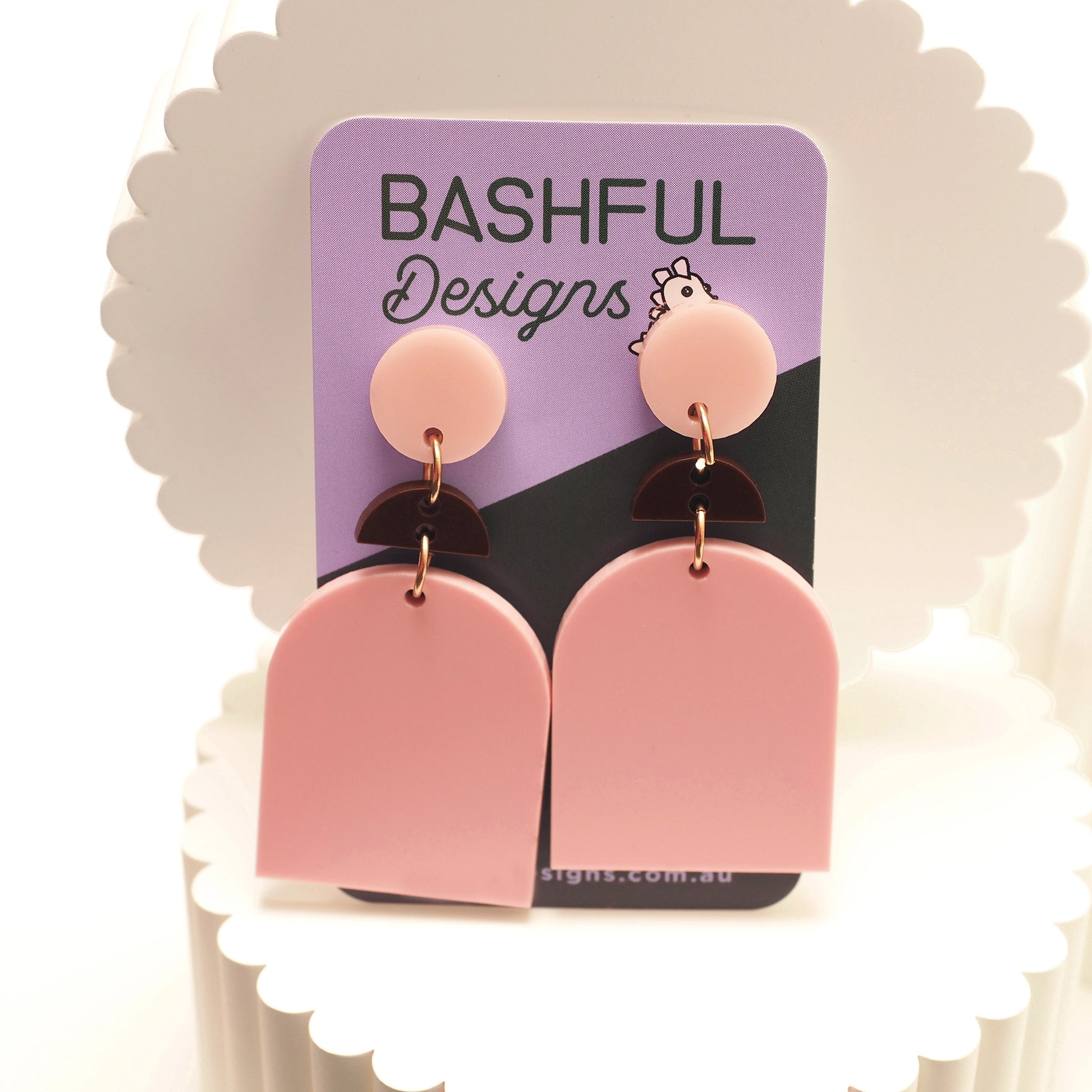 Light pink and chocolate brown acrylic earrings. They begin with a pale pink, round earring topper, then a chocolate brown half-moon shape, finishing with a pale pink solid archway. The earrings are shown on a 'Bashful Designs' earring card.