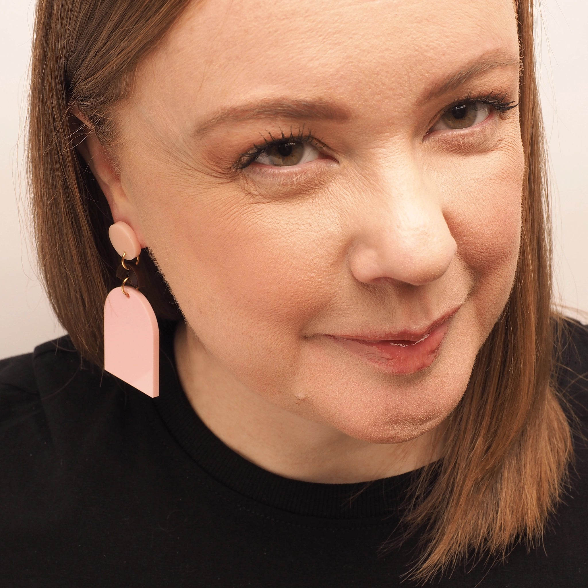 A curvy woman with red hair wearing a pair of light pink earrings in the shape of an archway. They feature a small chocolate brown half-moon shape above the arch and a light pink, round earring topper.