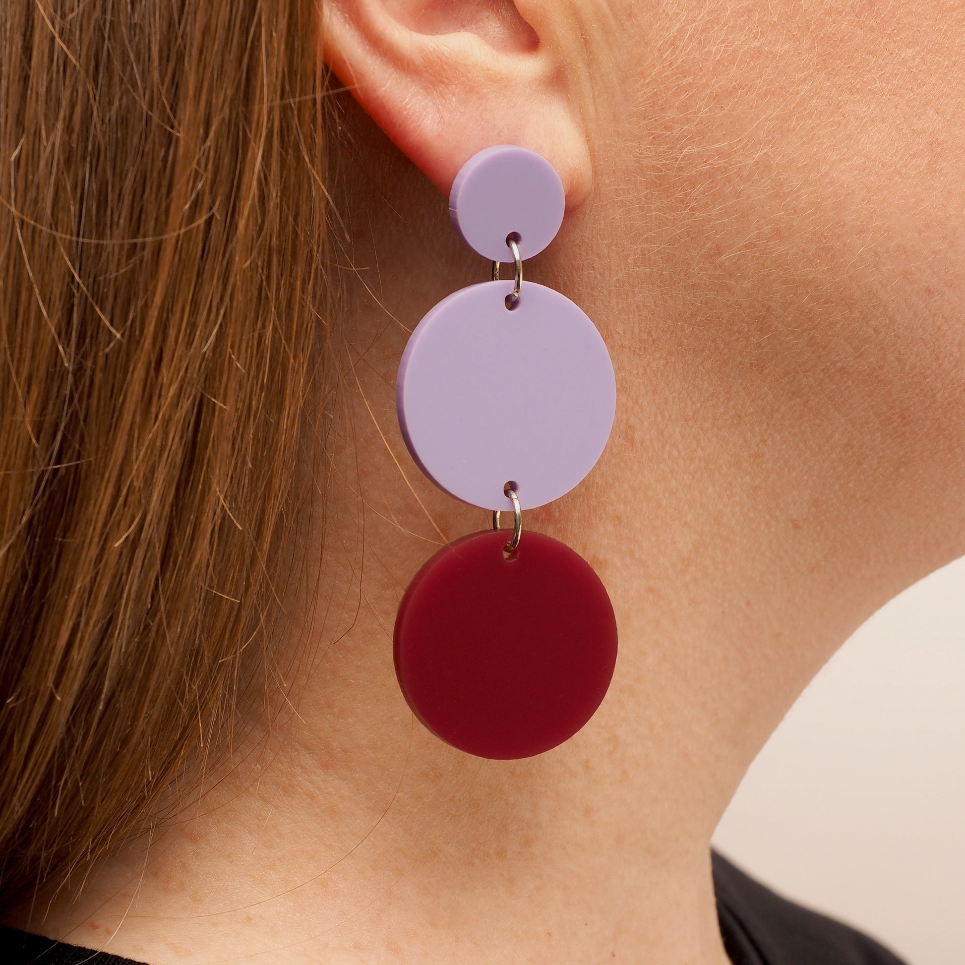 A person with straight red hair, wearing a pair of lilac and berry chandelier earrings. The earrings consist of three acrylic circles; the first is a small lilac circle, the second is a medium lilac circle, and the third is a medium berry coloured circle.
