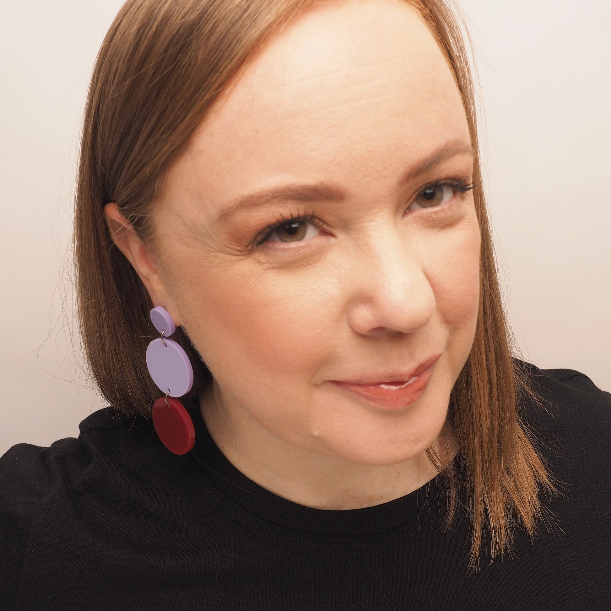 A curvy woman with straight red hair, wearing a pair of lilac and berry chandelier earrings. The earrings consist of three acrylic circles; the first is a small lilac circle, the second is a medium lilac circle, and the third is a medium berry coloured circle.