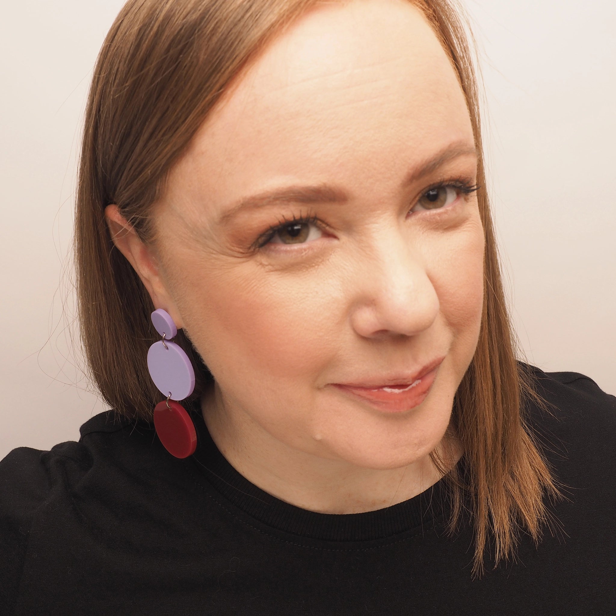 A curvy woman with straight red hair, wearing a pair of lilac and berry chandelier earrings. The earrings consist of three acrylic circles; the first is a small lilac circle, the second is a medium lilac circle, and the third is a medium berry coloured circle.
