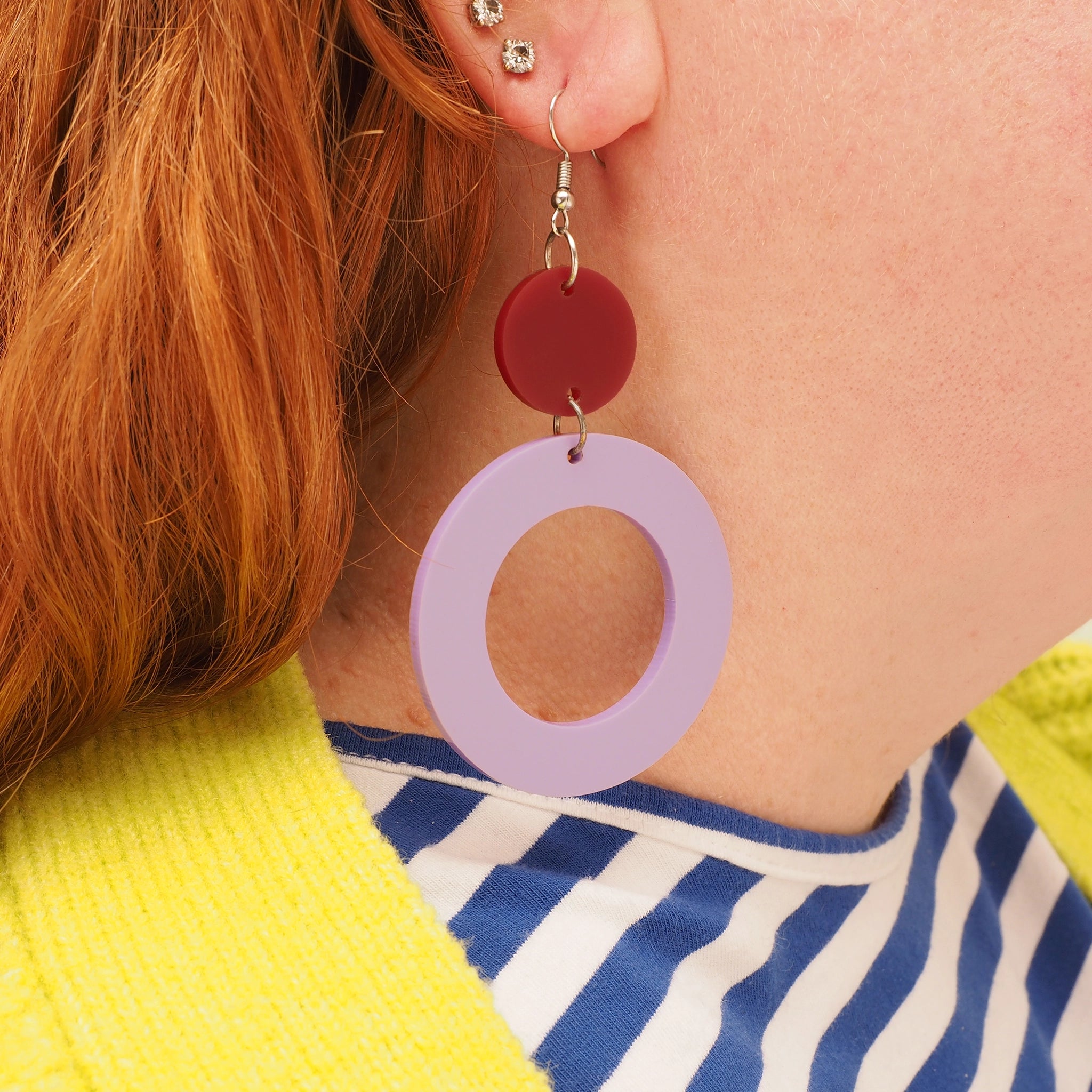 A person with straight red hair, wearing lilac and berry acrylic hoop-shaped dangles. These earrings are made of a small, dark berry coloured acrylic solid round atop a large, lilac acrylic hoop.