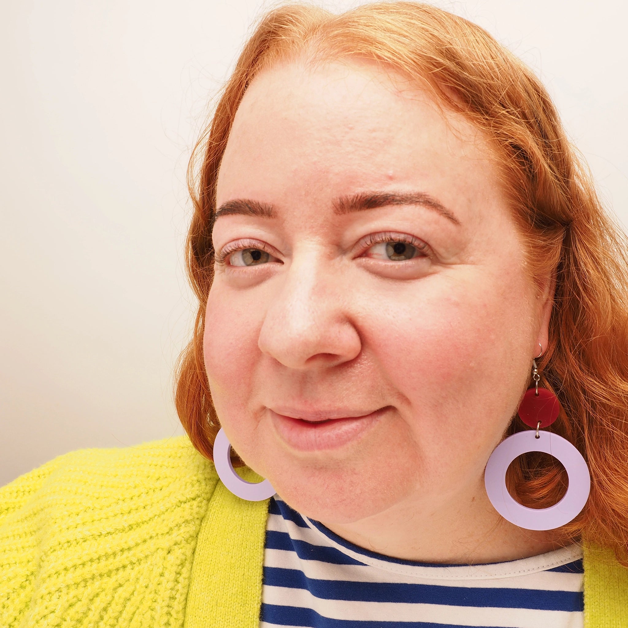 A curvy woman with straight red hair wearing lilac and berry acrylic hoop-shaped dangles. These earrings are made of a small, dark berry coloured acrylic solid round atop a large, lilac acrylic hoop.