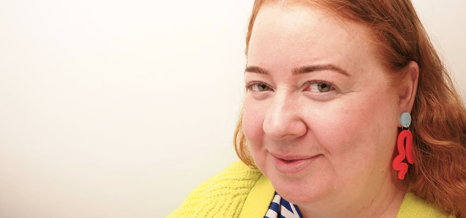 A curvy, red-headed model wearing The Twerk dangles. The earrings are made with a light blue acrylic button and a neon red acrylic squiggle in the shape of a body twerking. The pieces are connected by silver, surgical stainless steel jump rings.