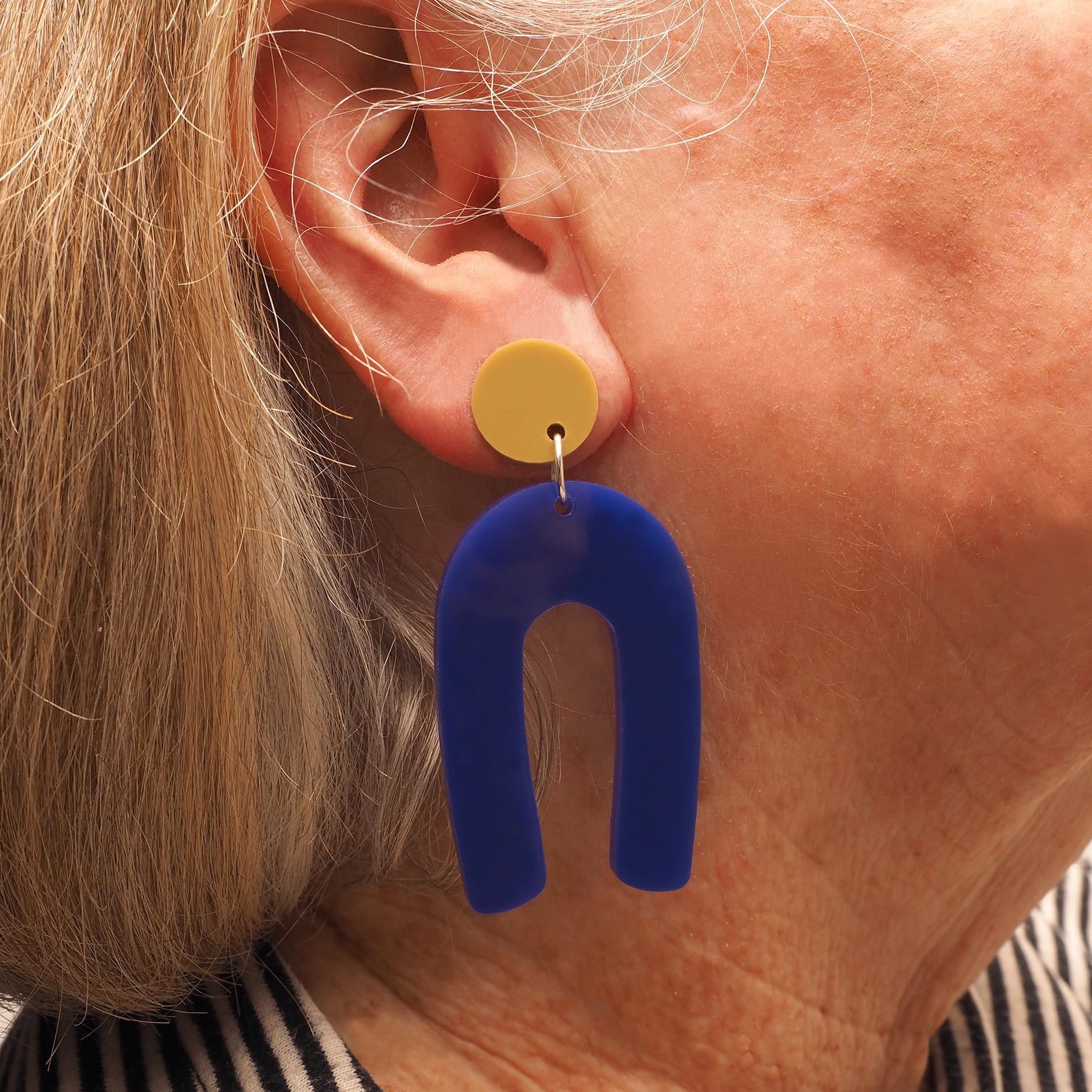 A woman with grey and blonde hair wearing navy blue acrylic earrings in the shape of an arch. These earrings hang from a light olive green earring topper.