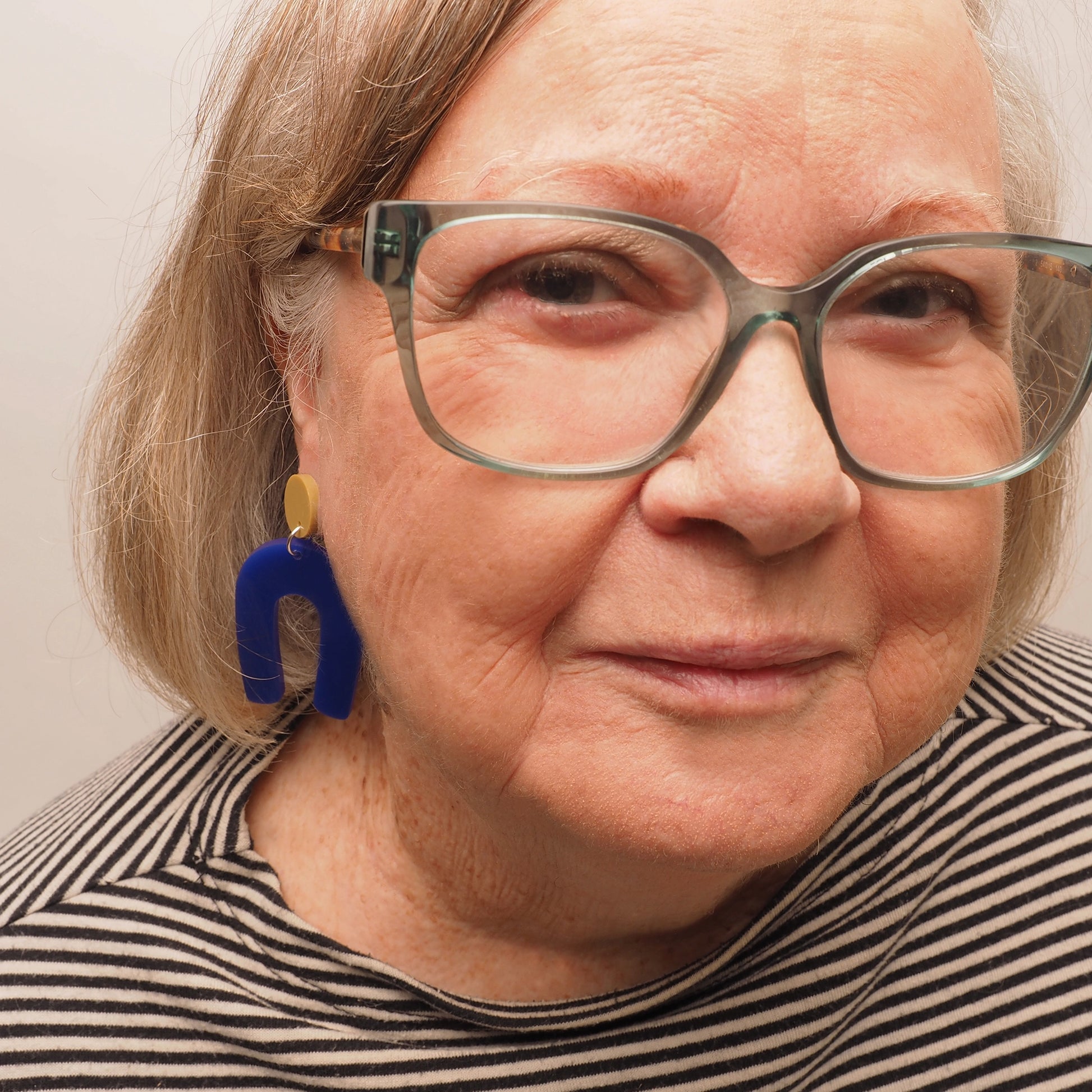 A woman in her 70's with grey and blonde hair wearing a pair of navy blue arch earrings with a light olive green round topper.