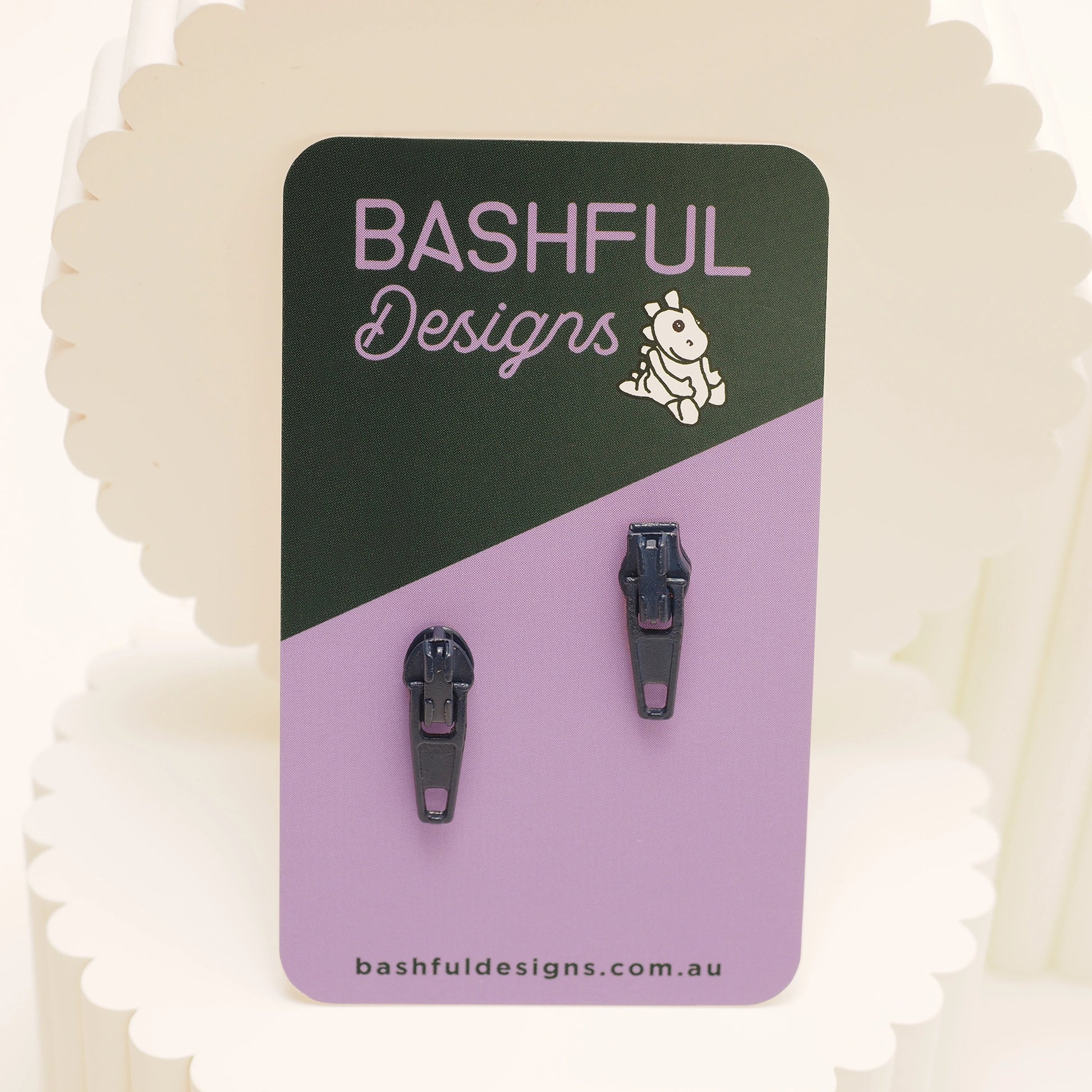 Dark blue zipper pull stud earrings on a 'Bashful Designs' branded card.